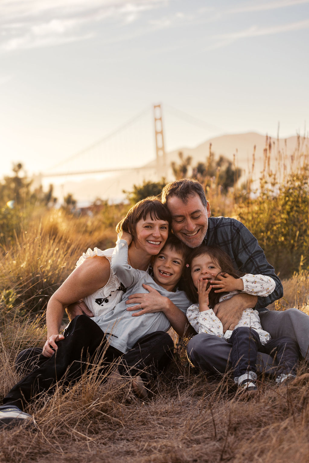 Family of four snuggled together in golden grass with the Golden Gate Bridge behind them at sunset