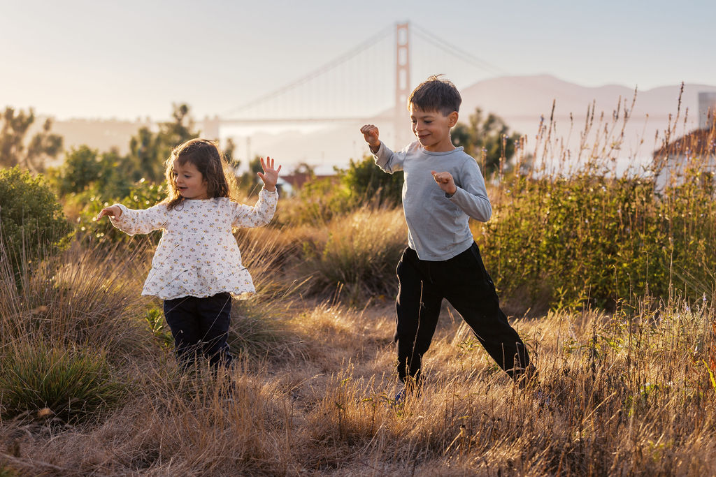 Two kids dancing in golden grass with the Golden Gate Bridge behind them at sunset, near Crissy Field in San Francisco