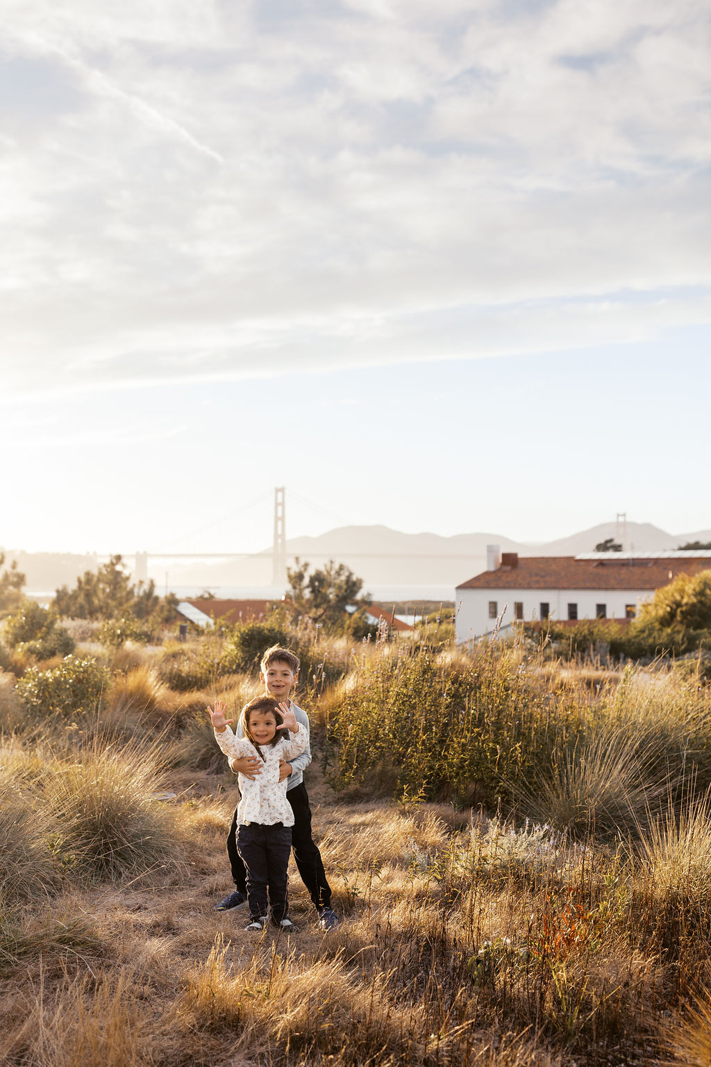 Two kids hugging in the grass under a big sky, the Golden Gate Bridge and a white building in the distance
