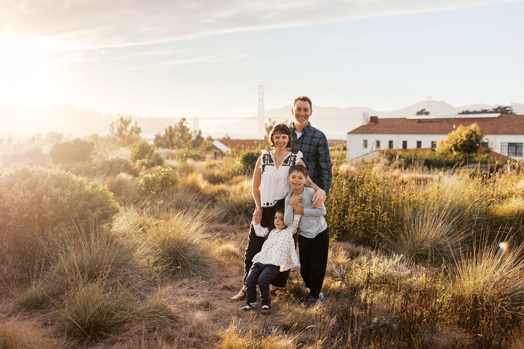 Family standing together in tall grass with the Golden Gate Bridge and a red-roofed building behind them at golden hour
