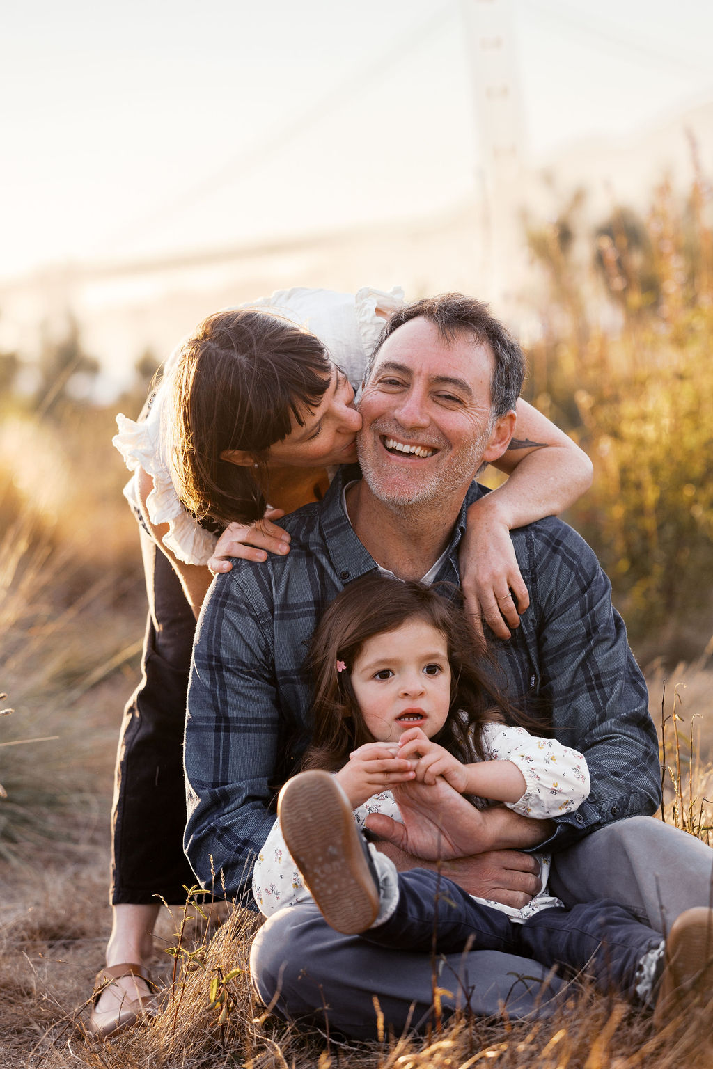 Mom kisses dad's cheek while their daughter sits in his lap, the Golden Gate Bridge soft in the background