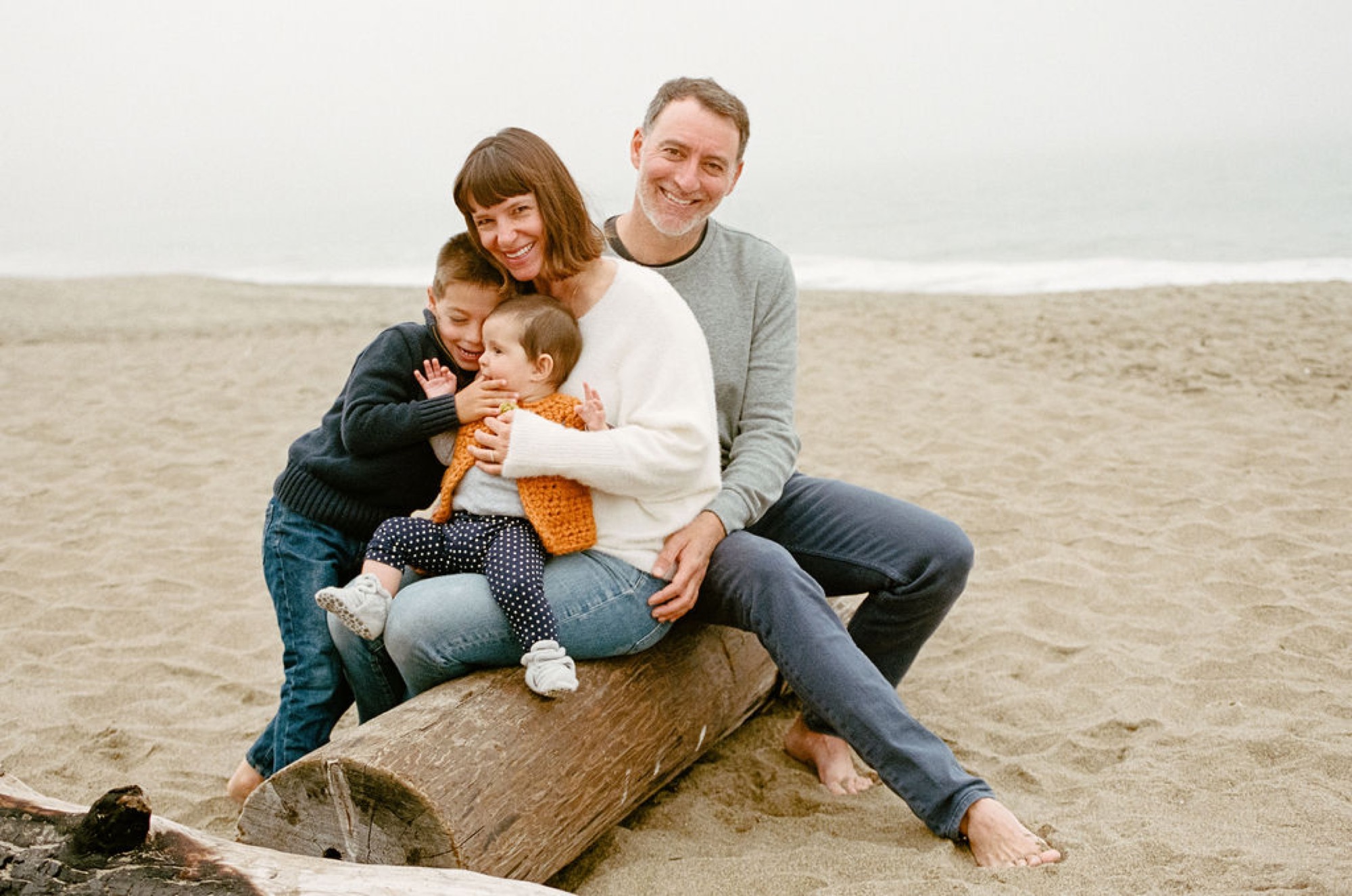 A family of four sits closely together on a weathered driftwood log, with soft overcast light illuminating their genuine smiles and relaxed embrace on the sandy beach