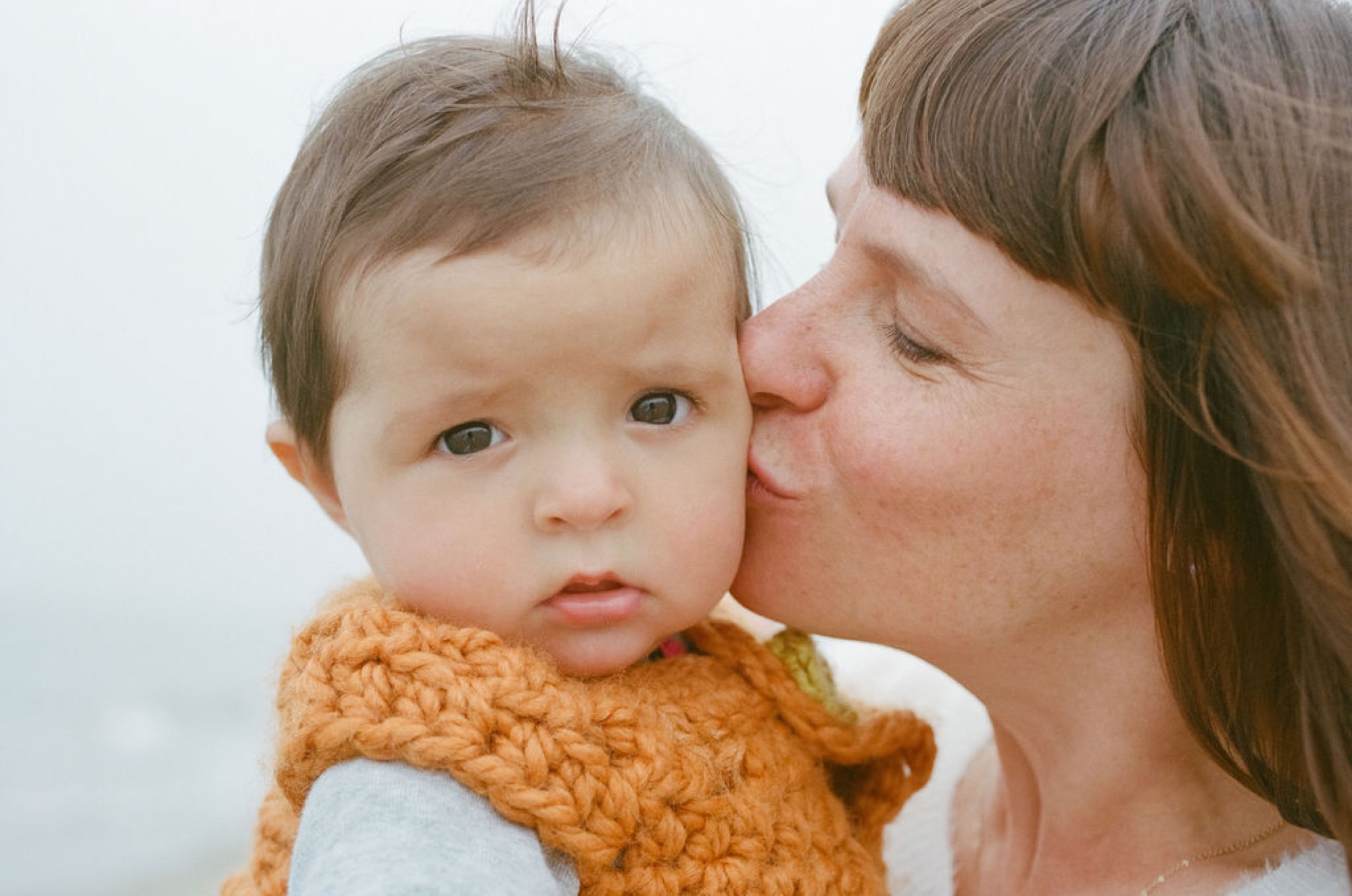 A mother kisses her baby girl's cheek while she holds a knitted orange toy, displaying serene contentment