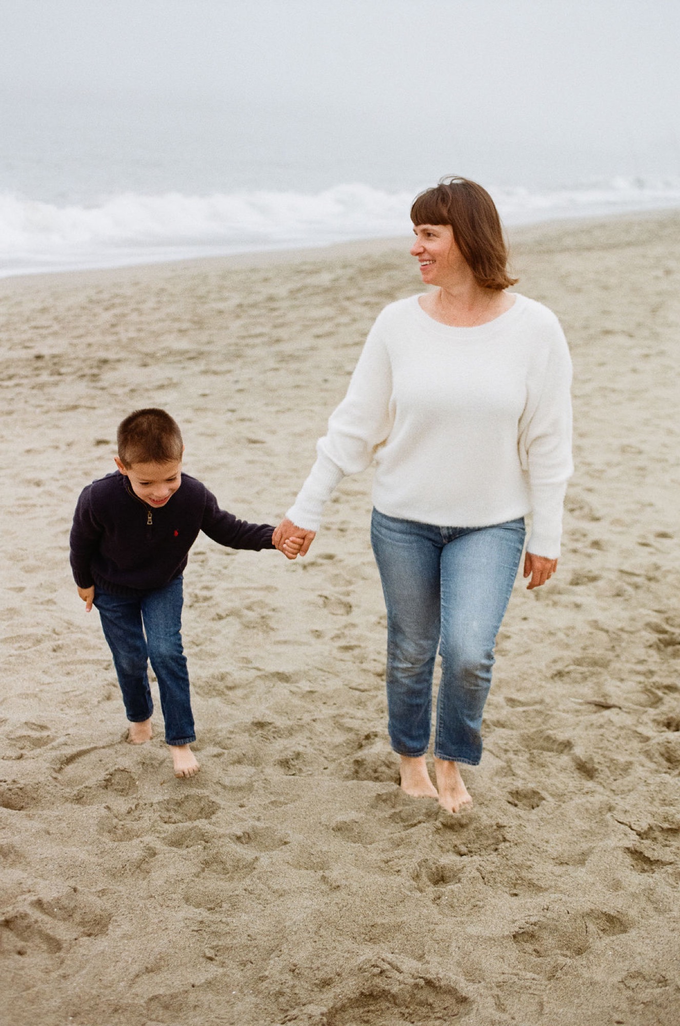 Woman and young child walk barefoot across sandy beach, holding hands while gazing toward misty ocean waves