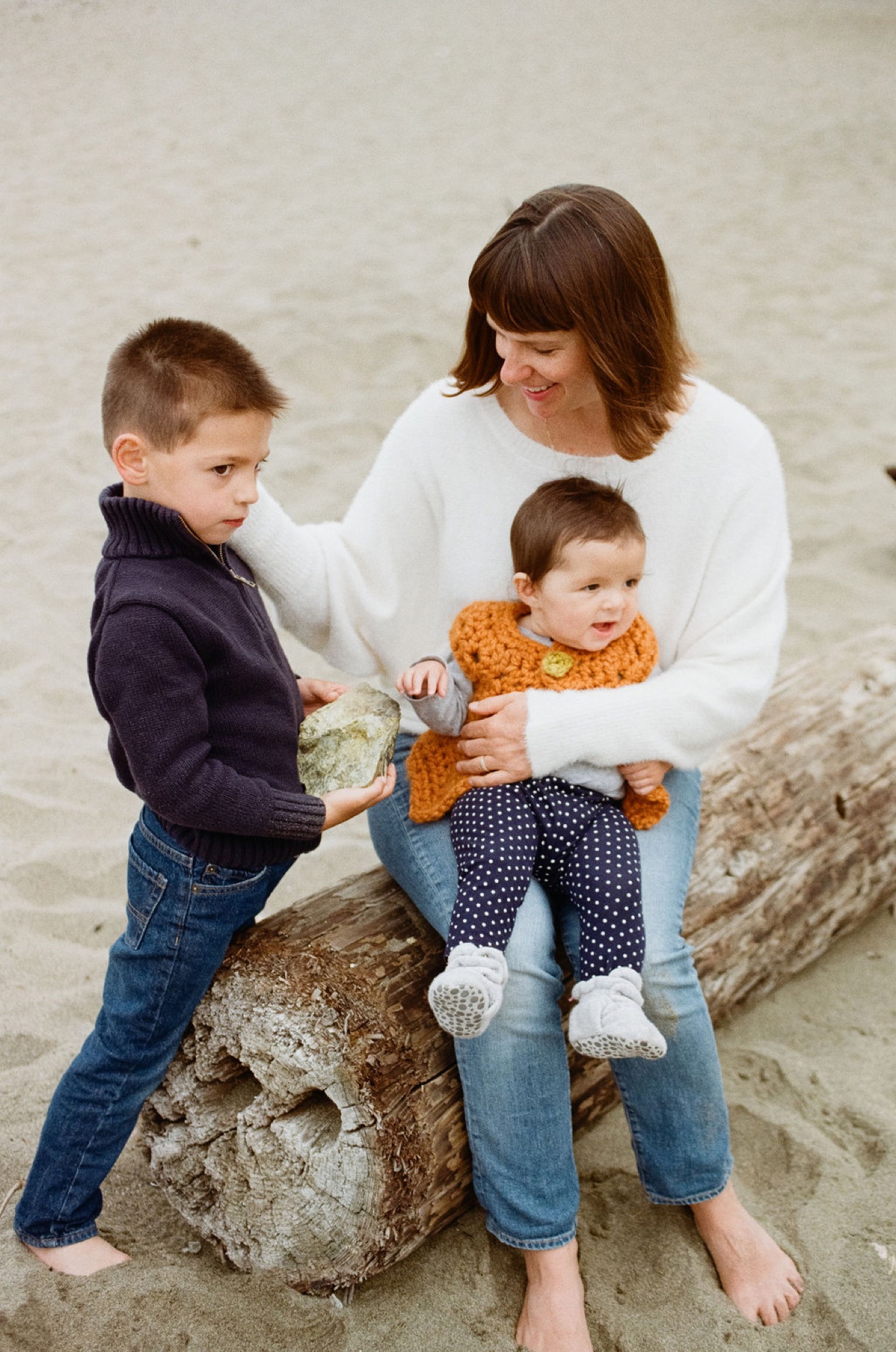 Woman and two young boys sit on a weathered log at the beach, the older boy holding a starfish while the younger child smiles in a yellow shirt