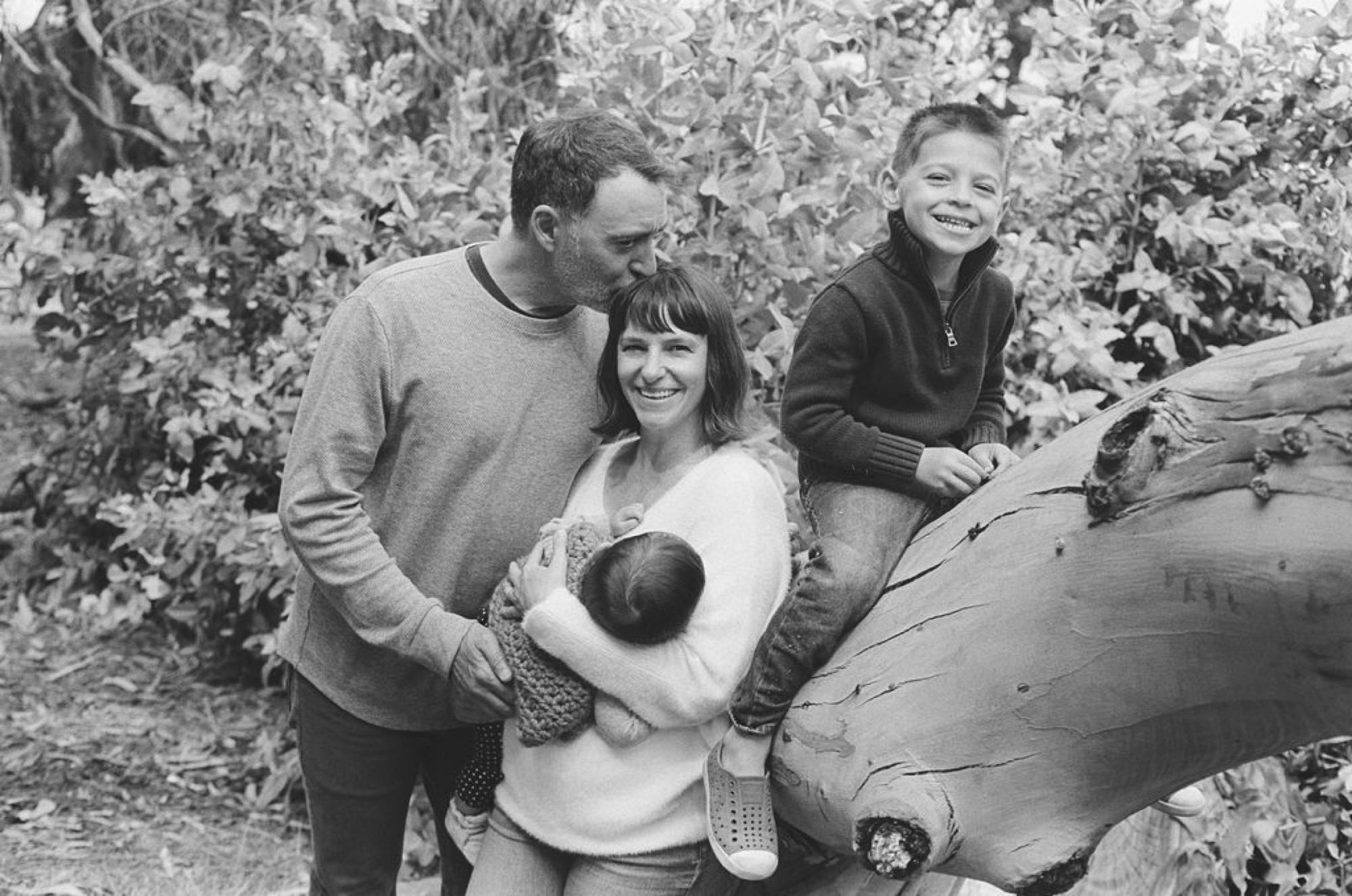 A man and woman smile warmly while their two children lean against a large weathered wooden post surrounded by dense coastal scrub vegetation