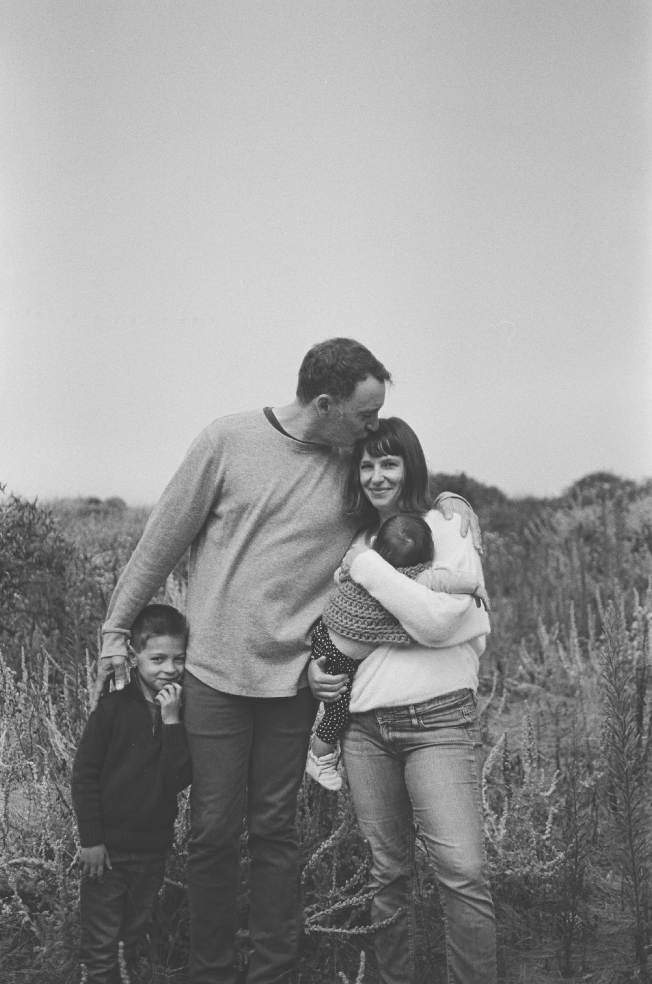 A father leans in to kiss his smiling wife while their young children stand close beside them in a windswept field