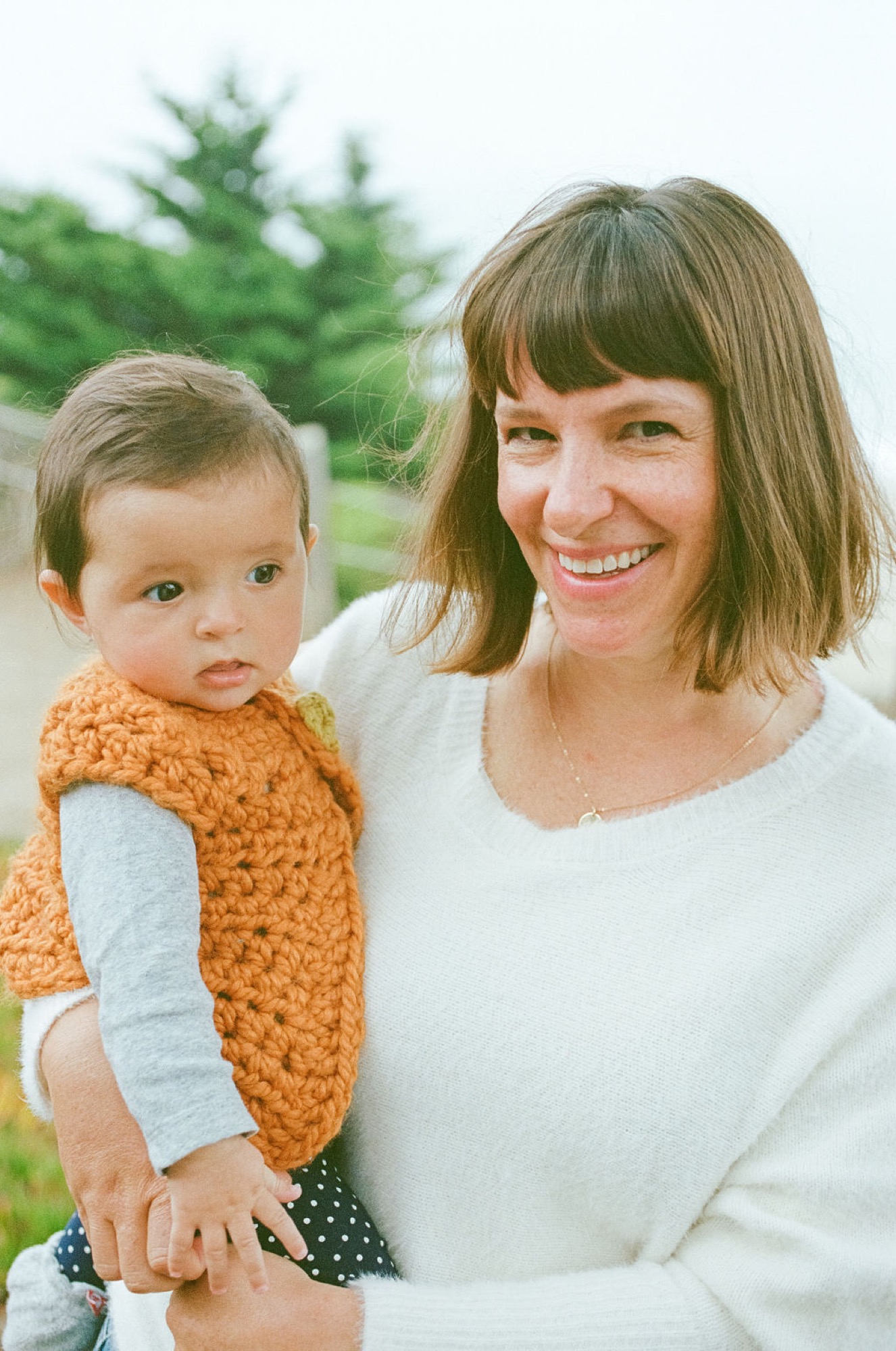 Woman in white shirt holds smiling toddler in orange sweater against soft green hillside backdrop