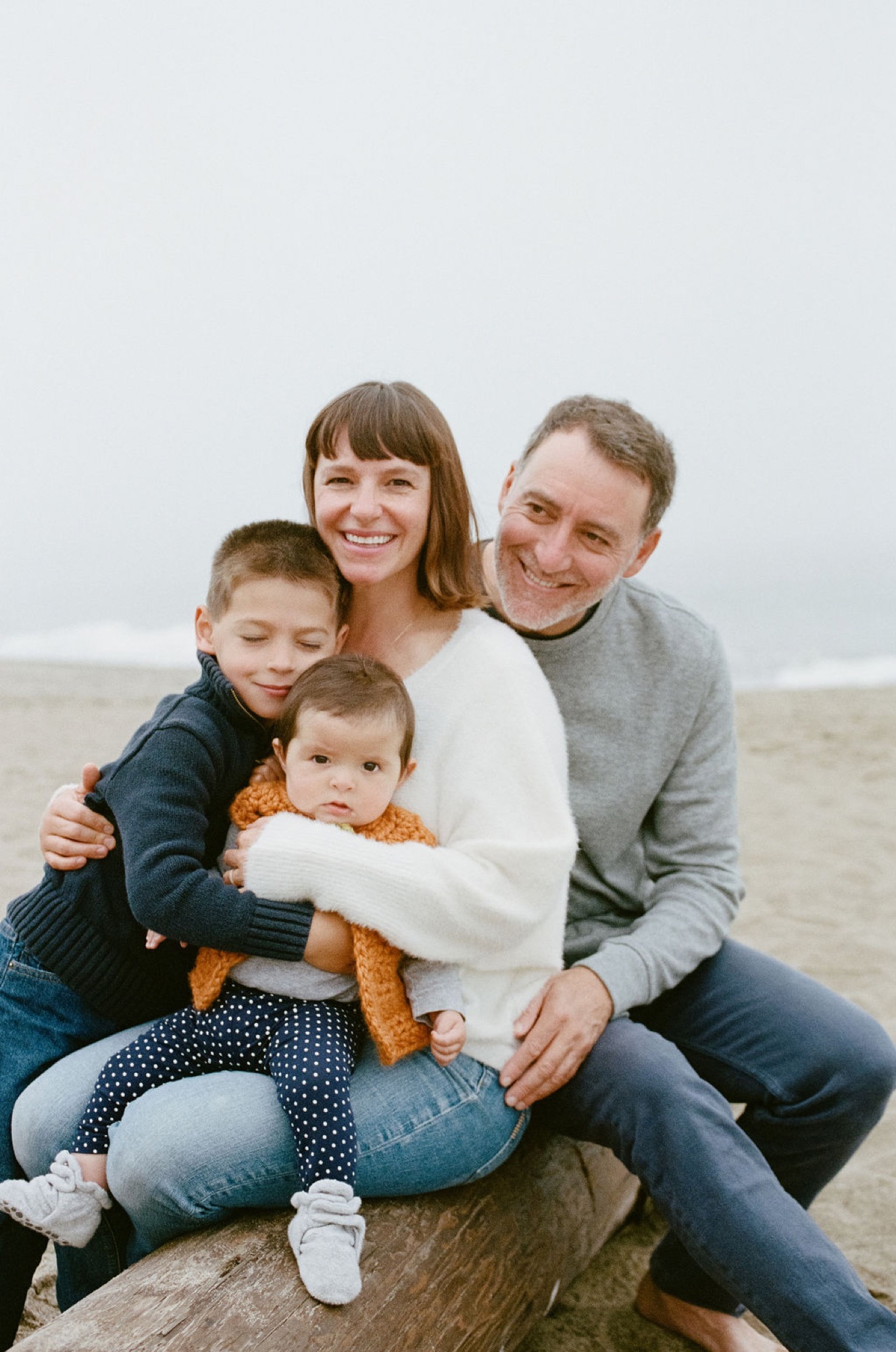 A family of four sits together on driftwood at the beach, smiling warmly at the camera with soft overcast light