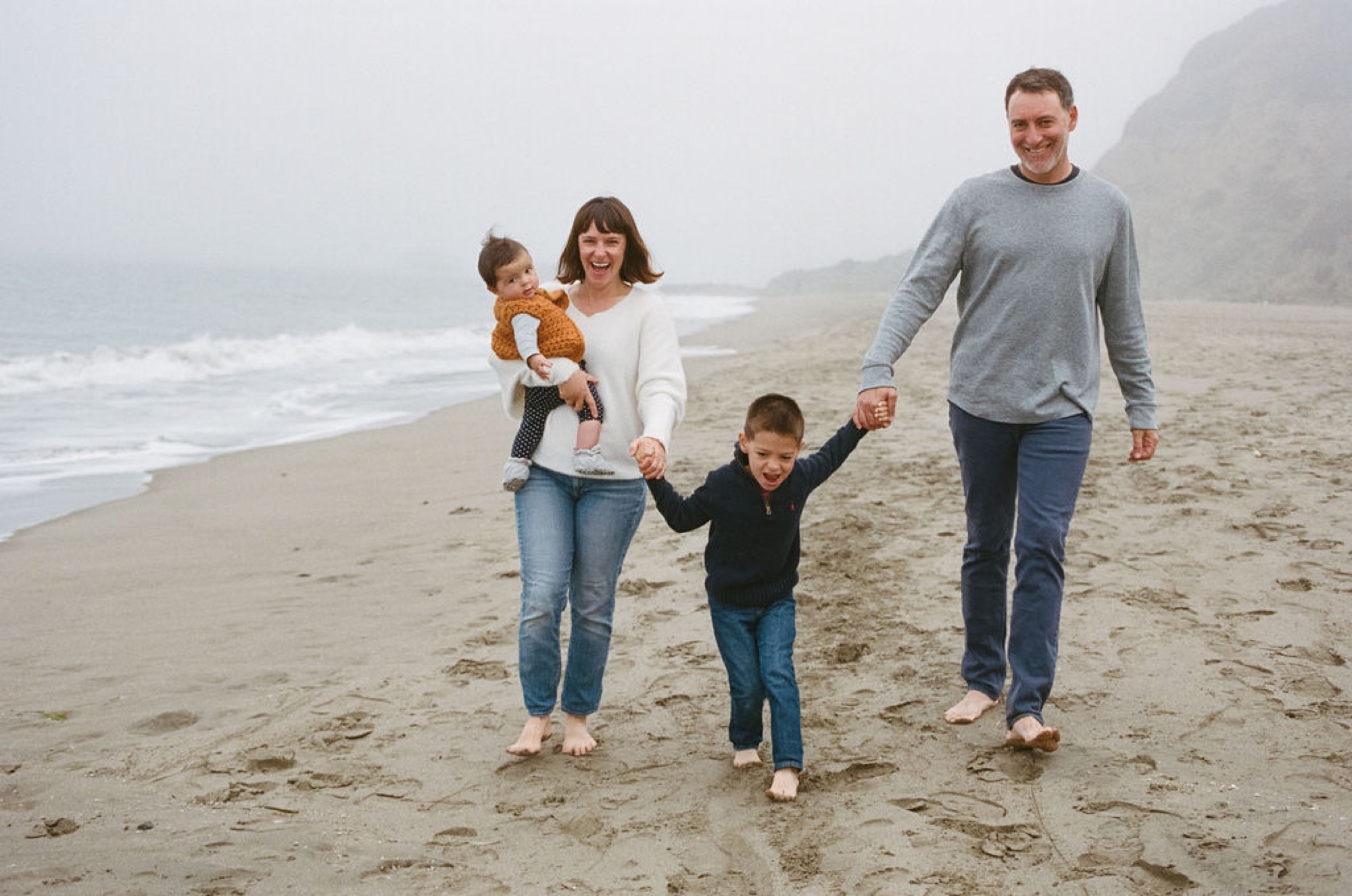 Family of four walks barefoot across misty Baker Beach sand, the youngest child lifted by mother while older boy holds father's hand