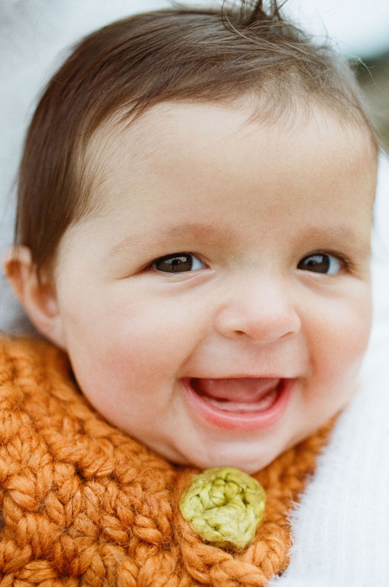 Laughing toddler in orange knit sweater with green button, bright eyes and open-mouthed smile radiating joy