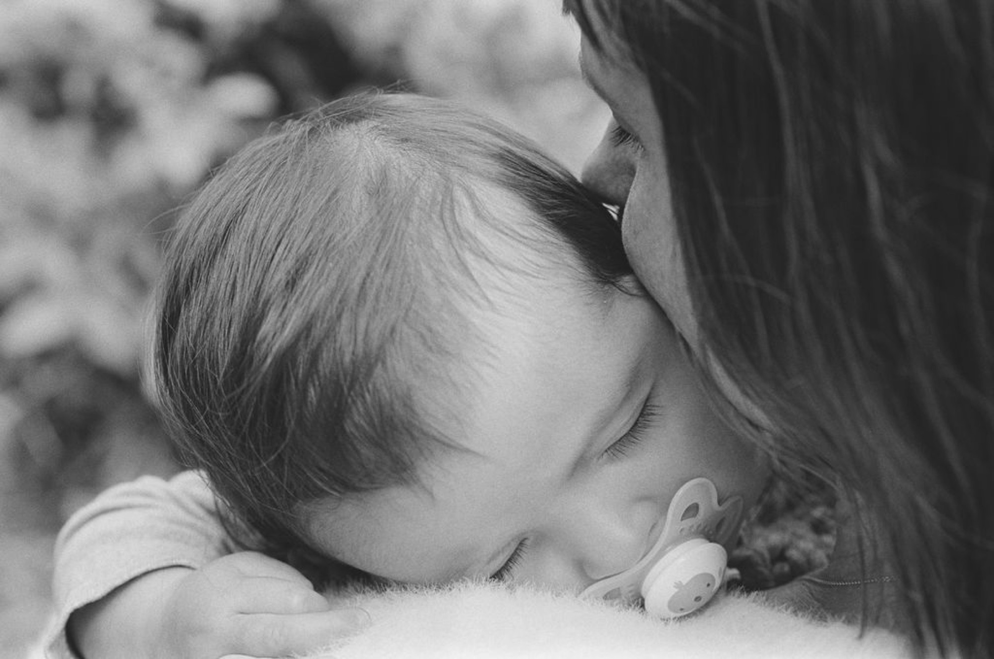 A young child rests peacefully against a woman's shoulder in soft, diffused black and white light