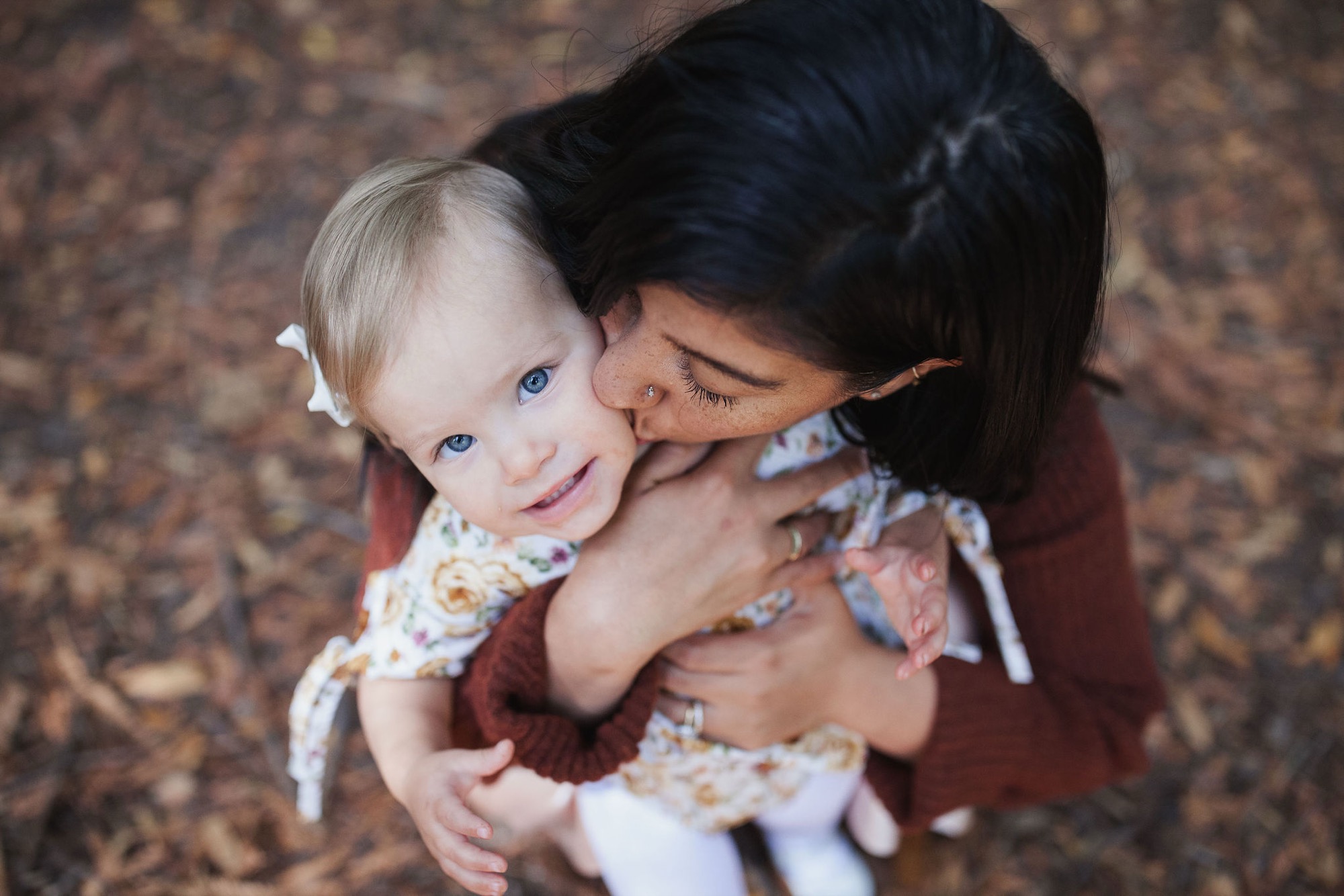 A woman holds a blonde toddler with striking blue eyes against a rust-colored ground, both gazing downward in tender connection