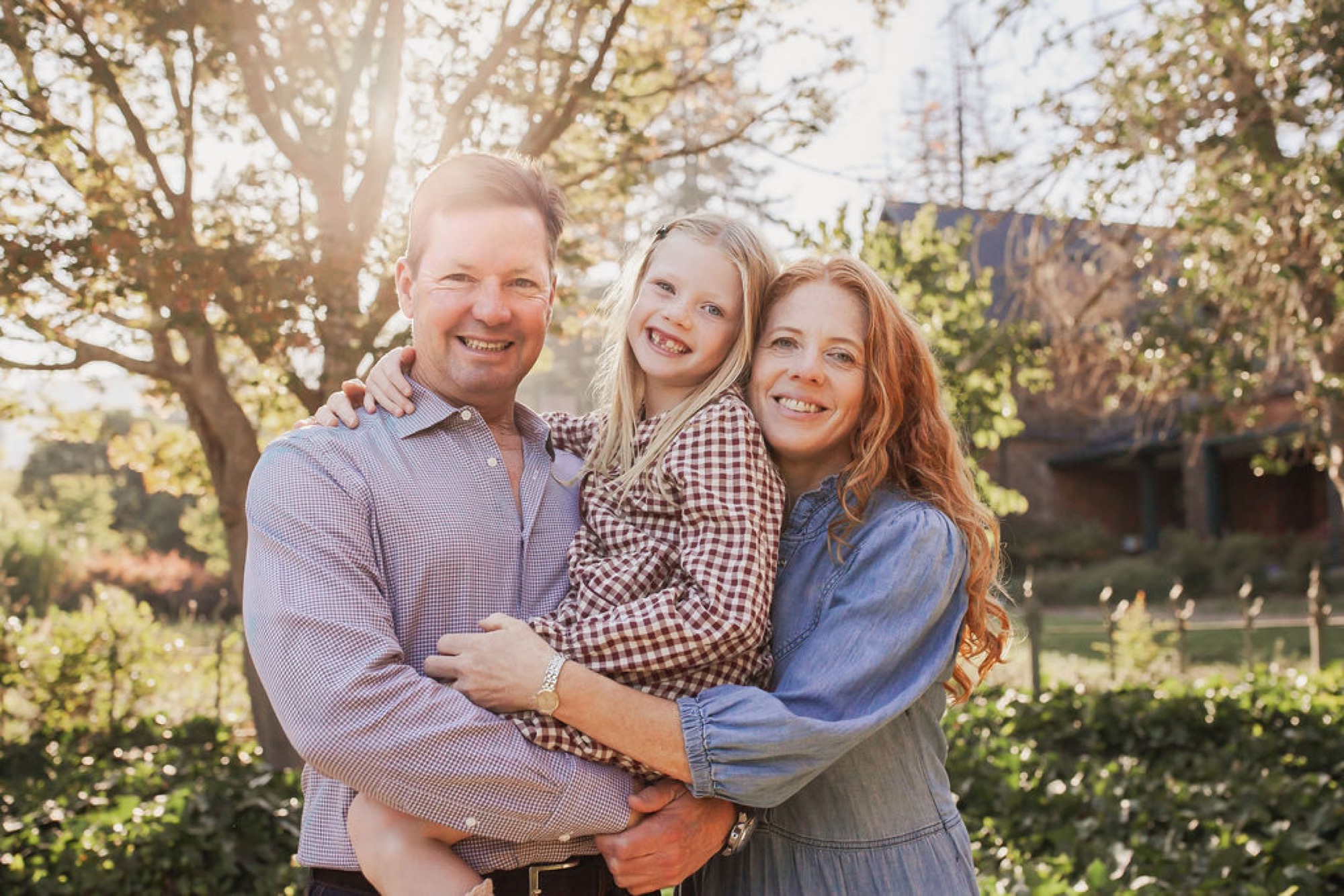 A smiling family of three stands together beneath golden-lit trees, with the father holding his young daughter while the mother embraces them both from the side
