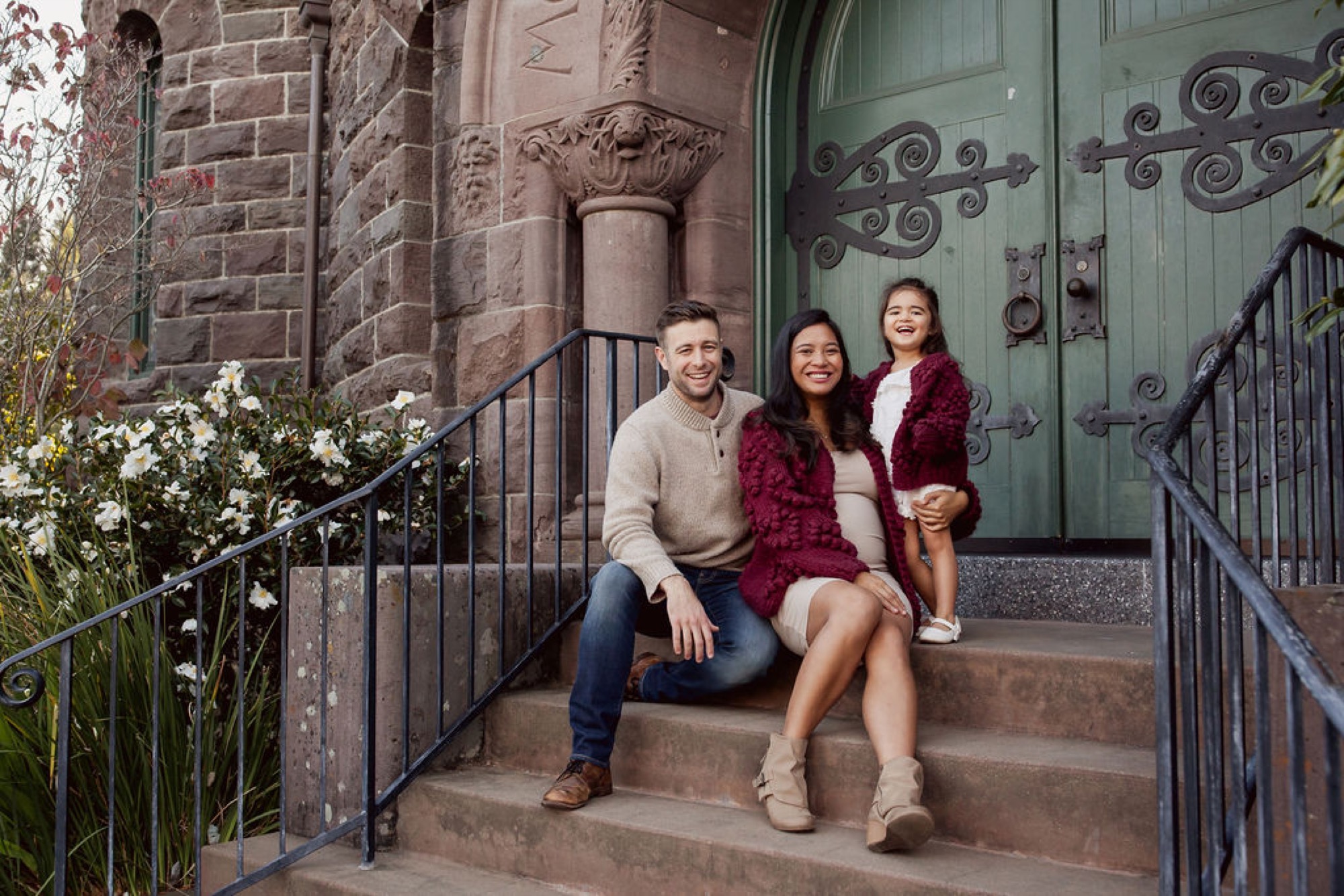 Three people sit closely on downtown stone steps beneath an ornate green door, wearing warm autumn tones and relaxed smiles — San Anselmo Family Photographer