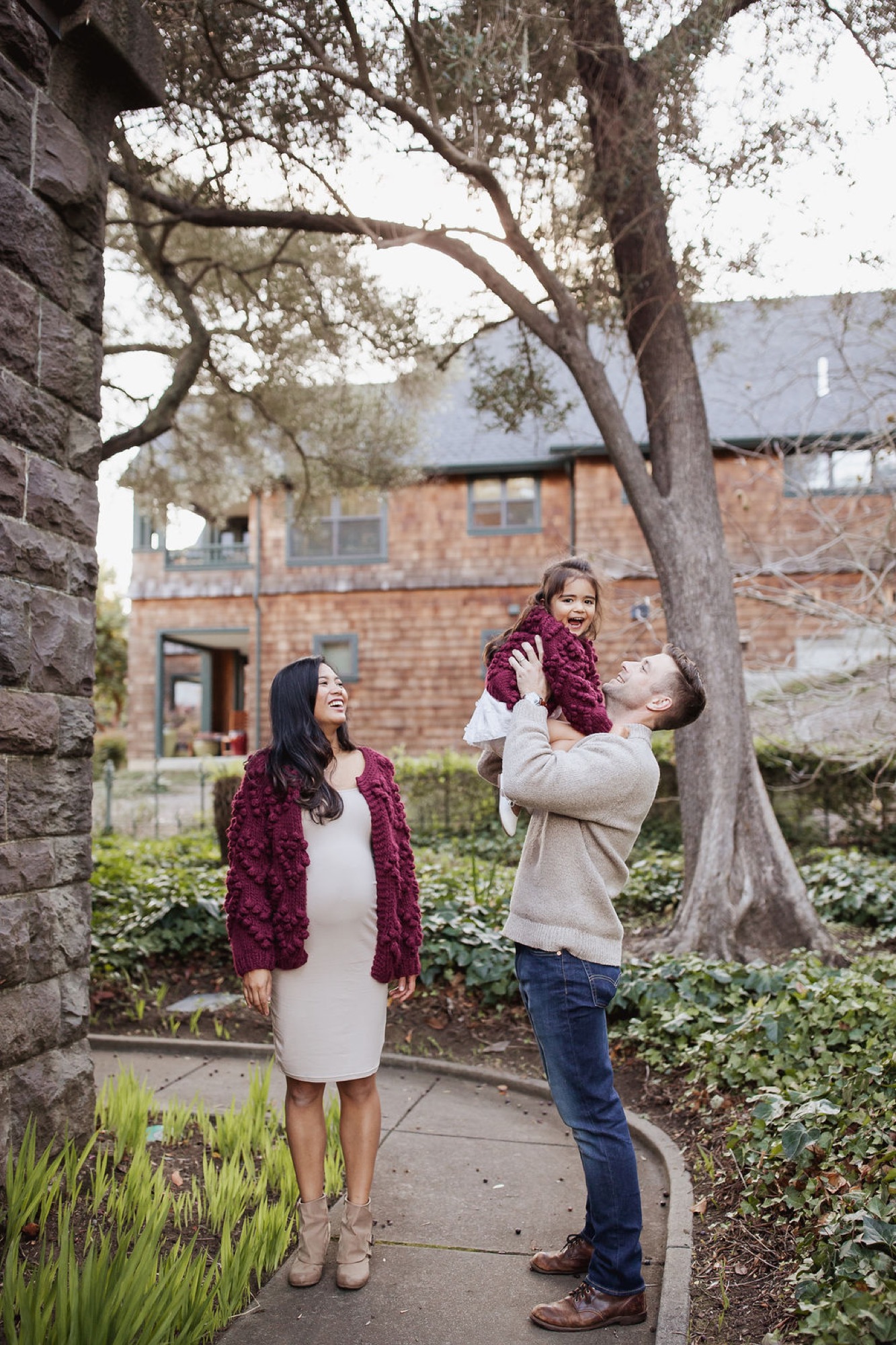 Parents and toddler stand beneath a sprawling tree on a garden pathway, the father lifting his child while the mother gazes upward in soft afternoon light — San Anselmo Family Photographer