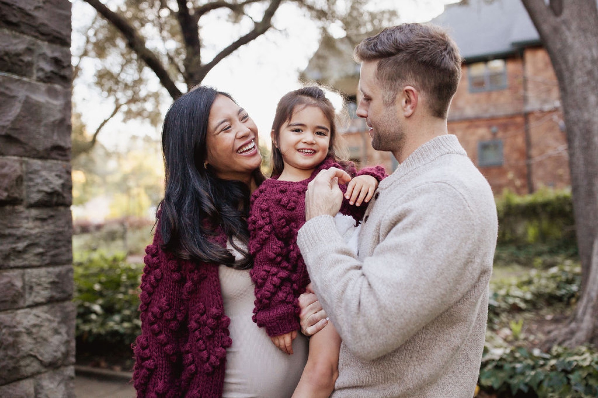 A family of three stands together between stone pillars, the child in burgundy between parents who gaze at each other warmly in dappled afternoon light — San Anselmo Family Photographer
