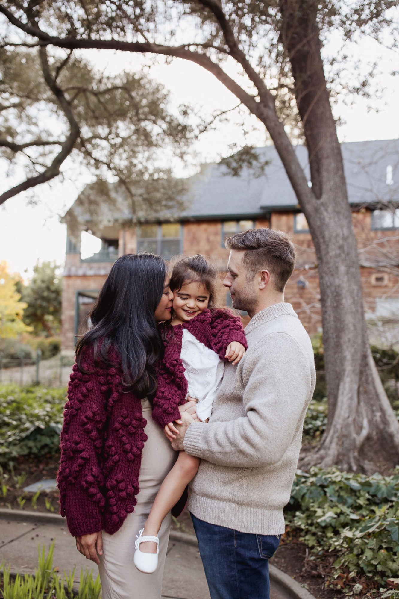 Parents hold their young daughter beneath a sprawling tree, sharing an intimate moment of laughter and connection in front of a brick home