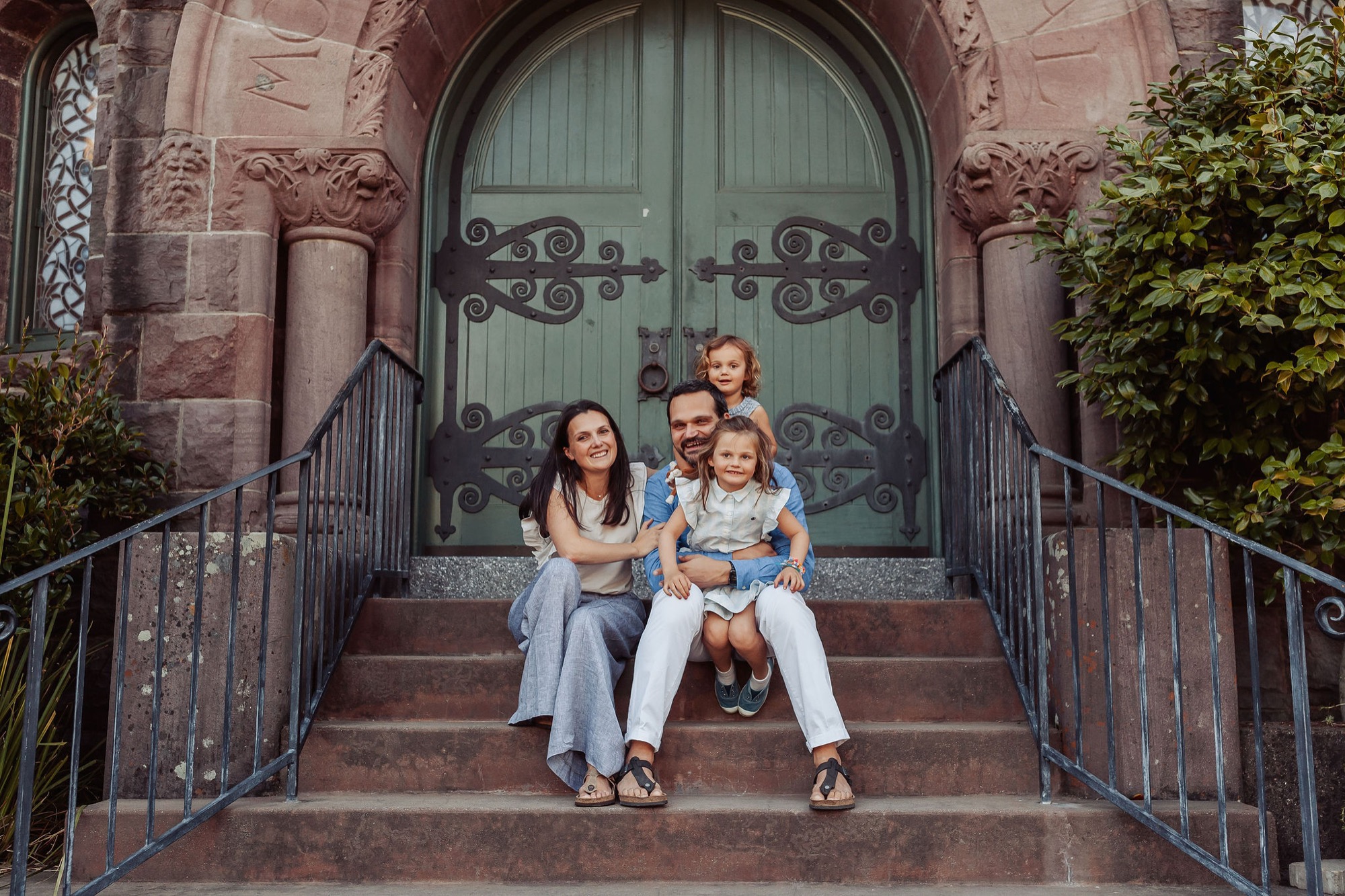 Family of four sits centered on brick steps before an ornate arched doorway, with the parents flanking two young children in casual seated poses