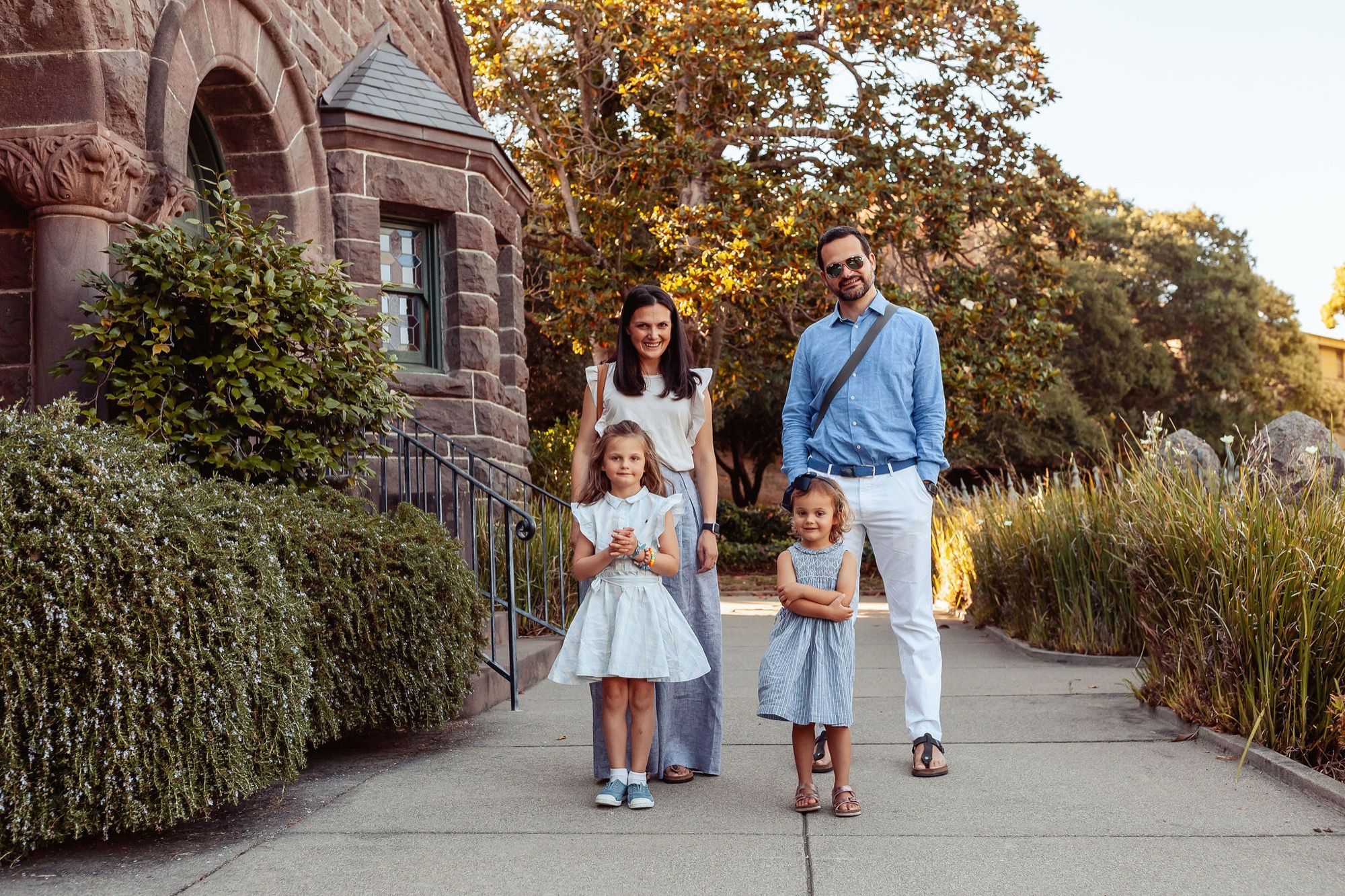 Family of four walks toward golden sunset along a tree-lined downtown pathway, the youngest child on a scooter while parents guide their daughters forward