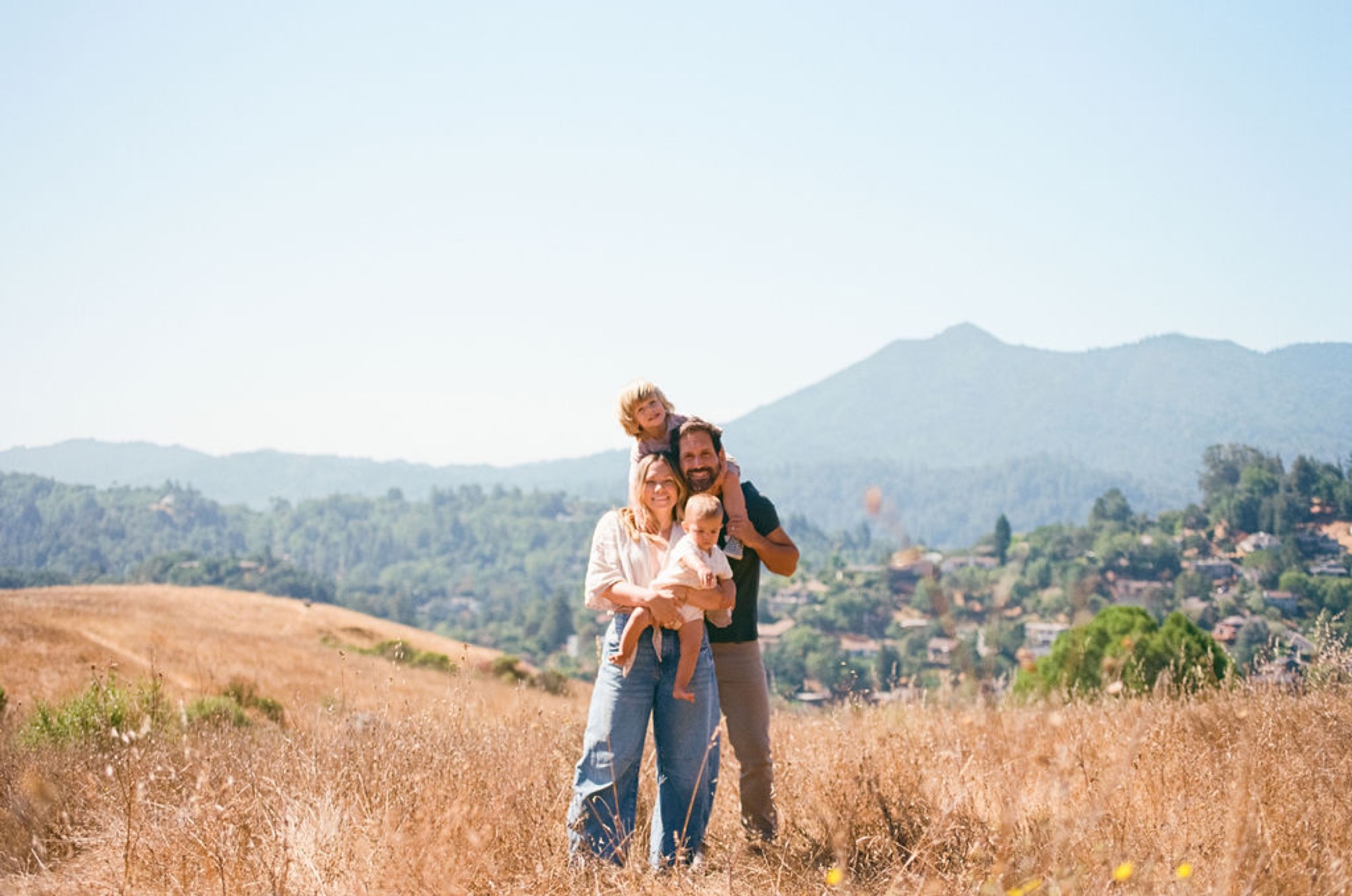 Father carries toddler on shoulders while mother holds infant, standing in golden hillside grass with forested mountains rising behind them