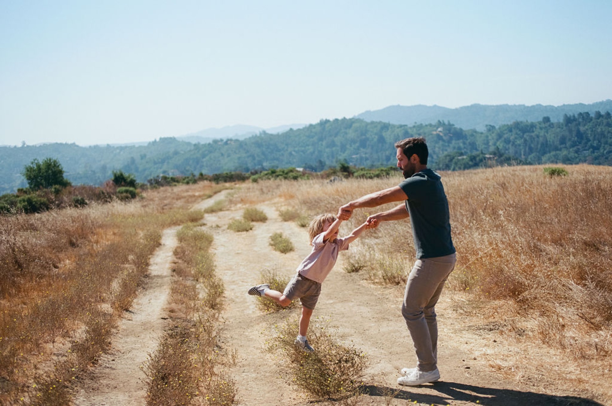 A father swings his young son by the arms along a dusty trail, both mid-motion with joy, golden hillsides and forested mountains stretching behind them