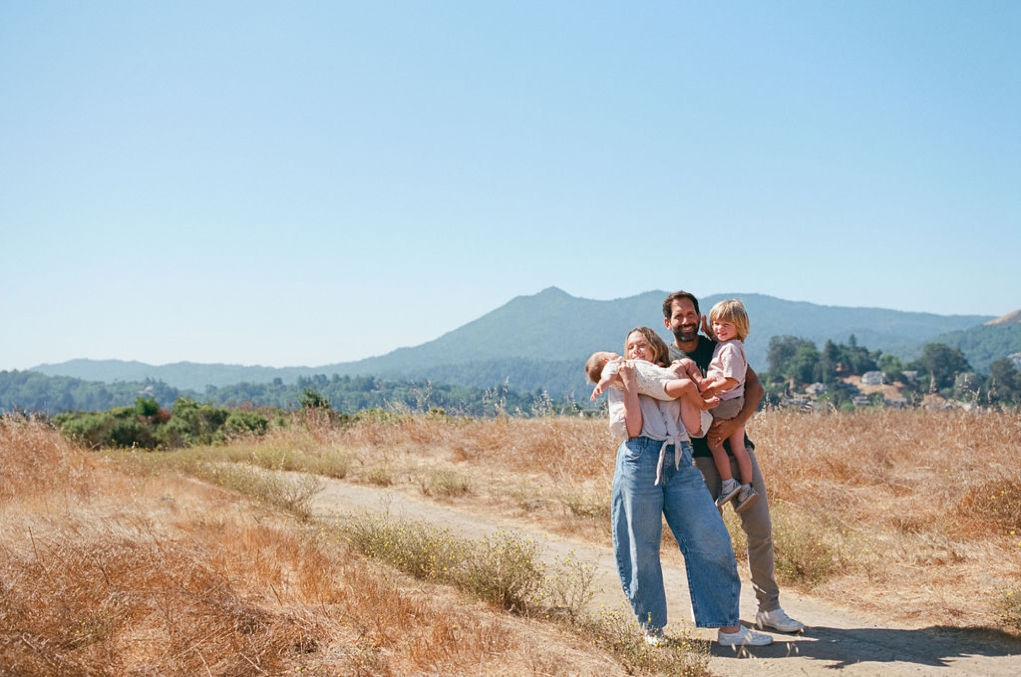 A father and mother hold their two young children in a dry grass field with rolling mountains rising behind them under clear sky