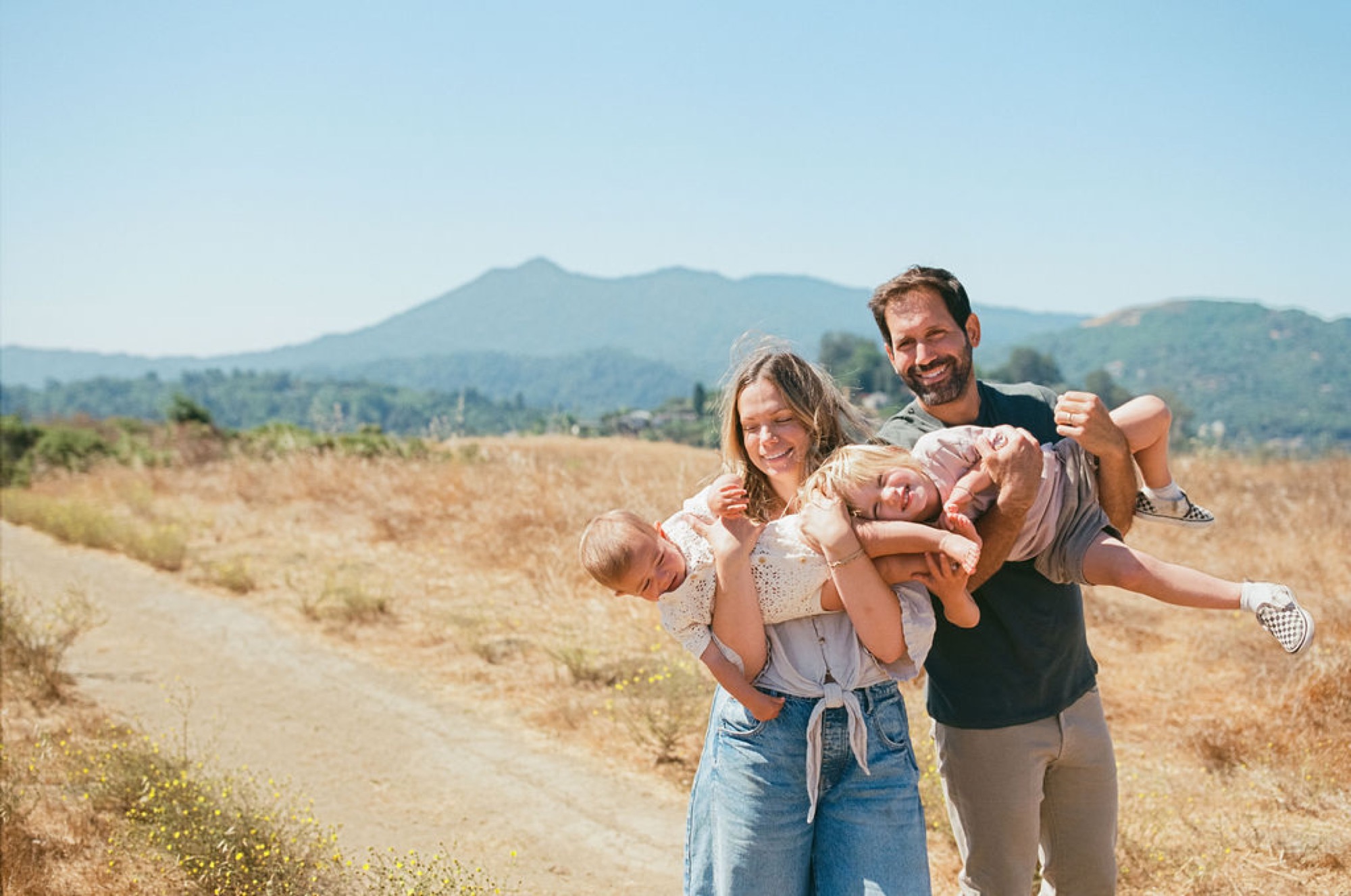 Parents beam while holding three young children aloft in a sun-drenched valley, mountains rising softly behind them