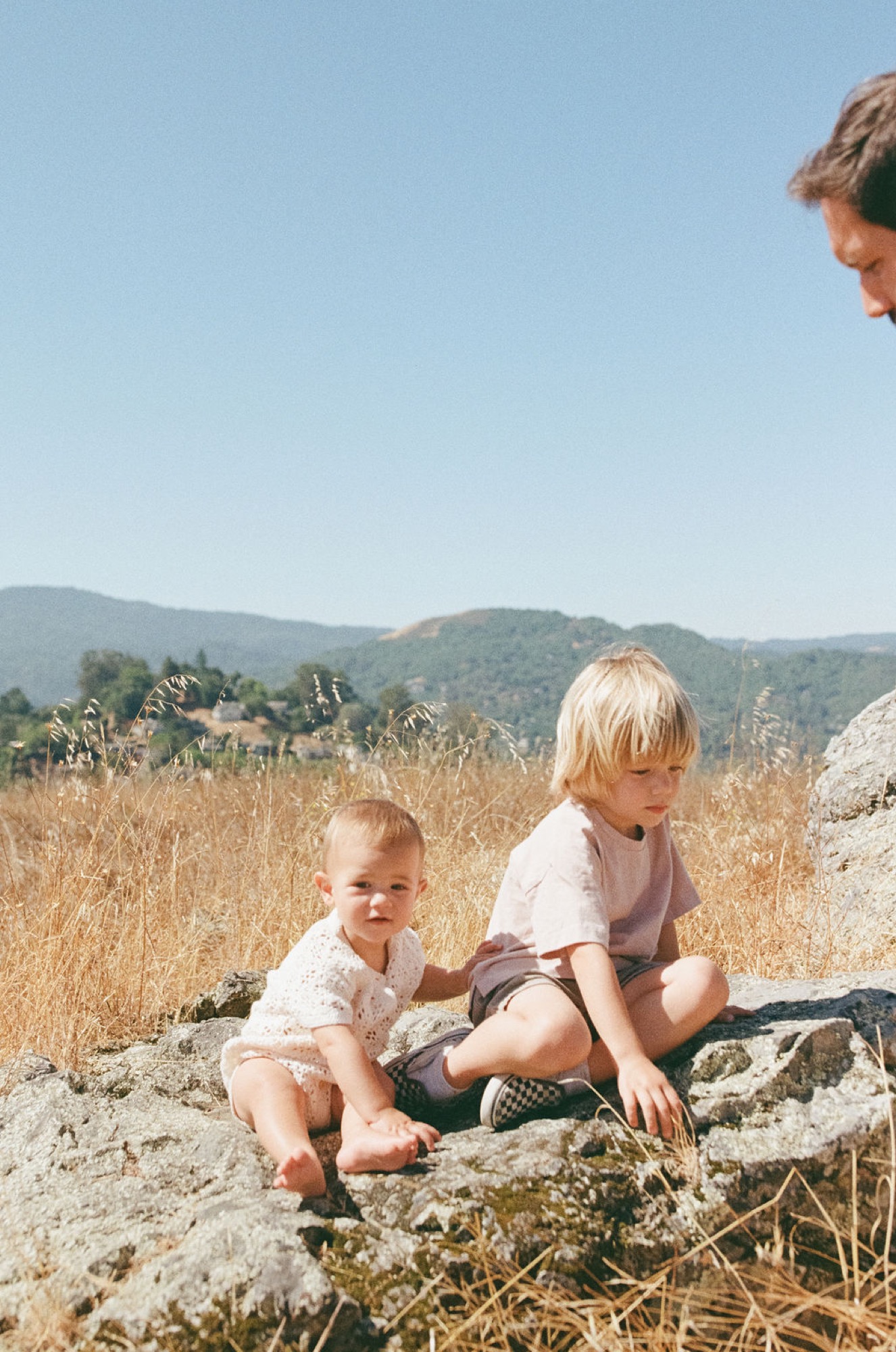 Two young children sit barefoot on weathered rocks overlooking golden grassland, with forested hills and clear sky beyond