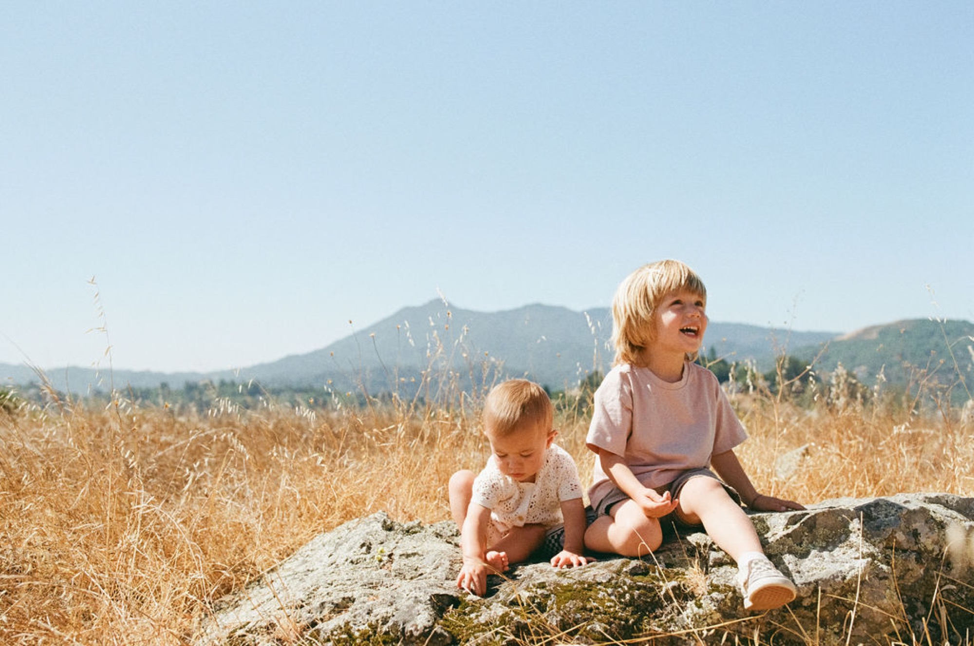 Two young children sit on a moss-covered boulder in golden dry grass, with mountains rising behind them under clear skies