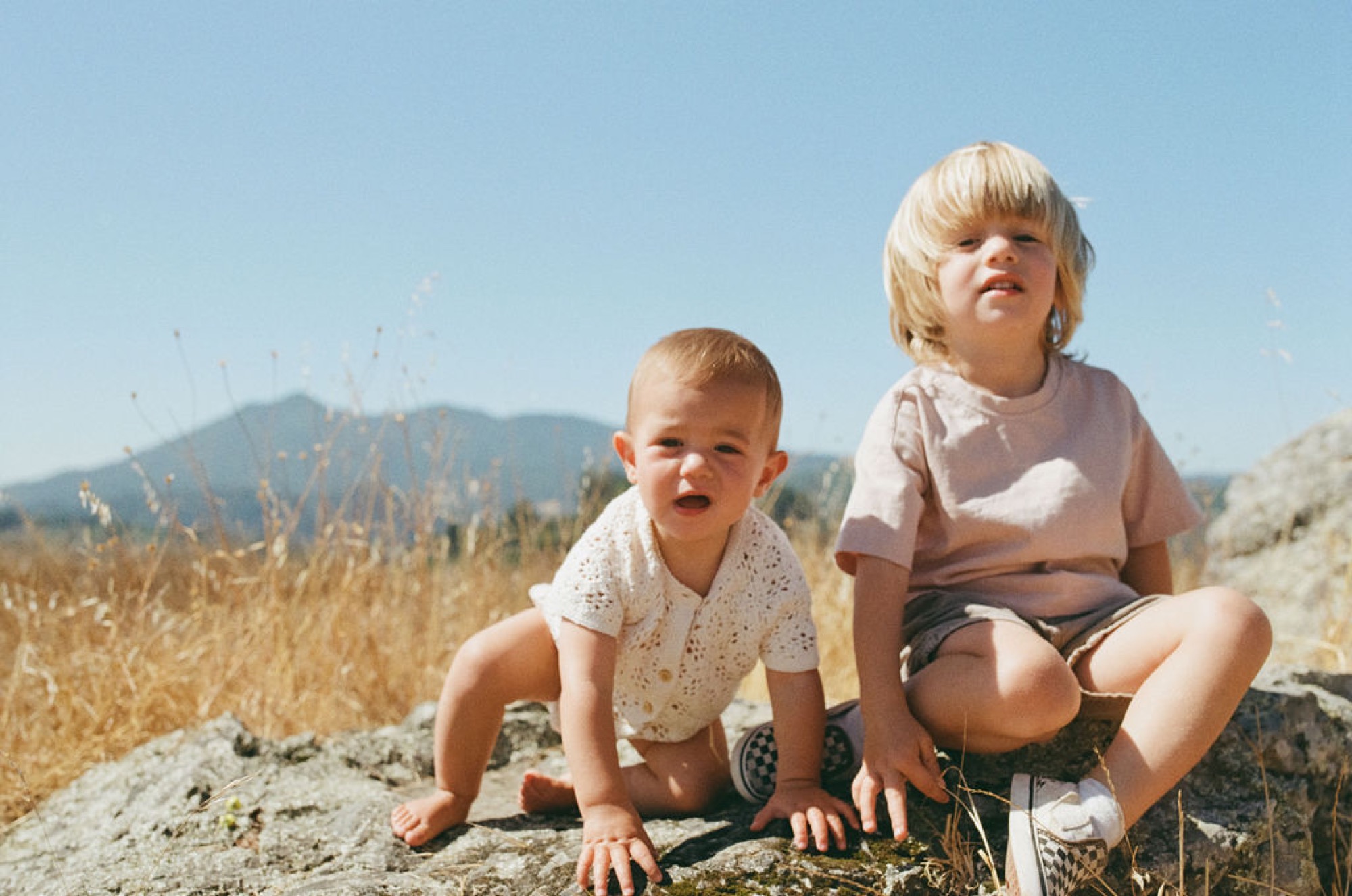 Two blonde toddlers crouch on sun-warmed rocks overlooking golden hillsides and distant mountains under clear blue sky