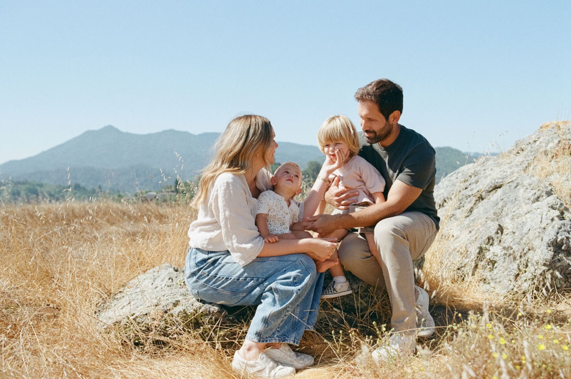 Parents crouch beside two young children on rocky terrain, bathed in warm afternoon light with mountains visible across the dry grassland