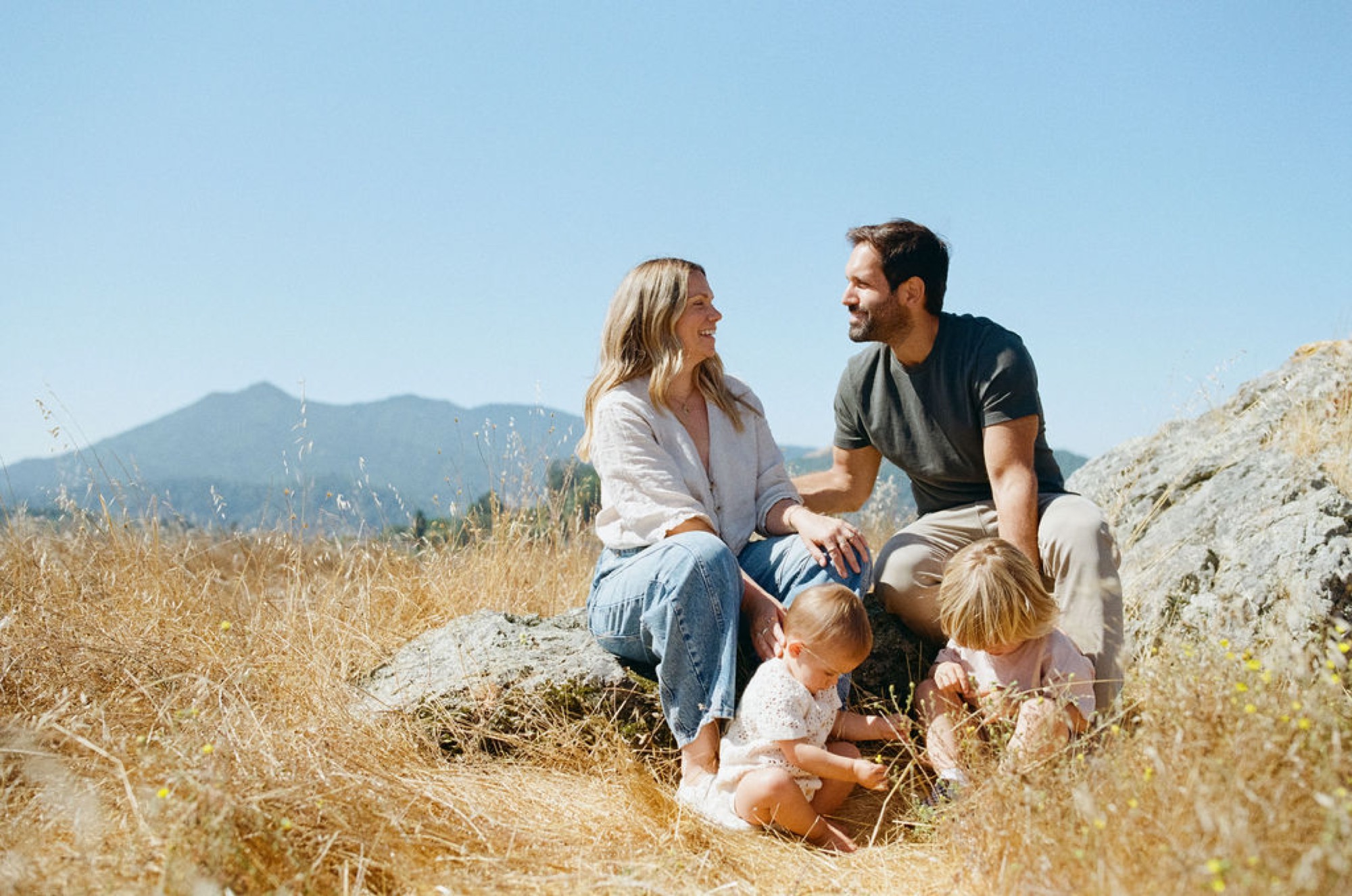 Parents and two young children sit together on rocky terrain, gazing at each other with warm smiles beneath clear blue skies and distant mountains
