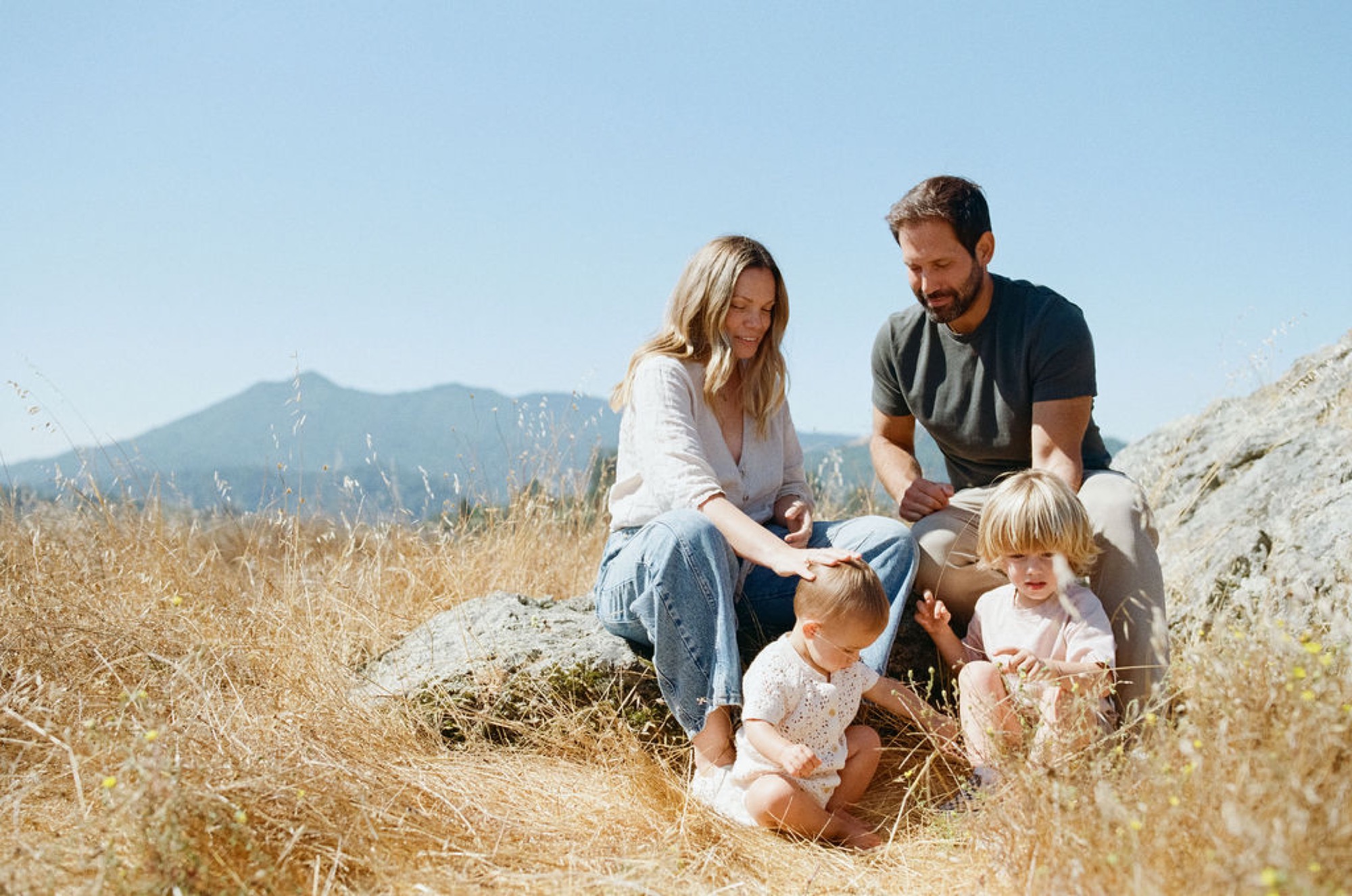 A family of four kneels together on sun-dried grass with a distant mountain peak framed behind them under clear blue sky
