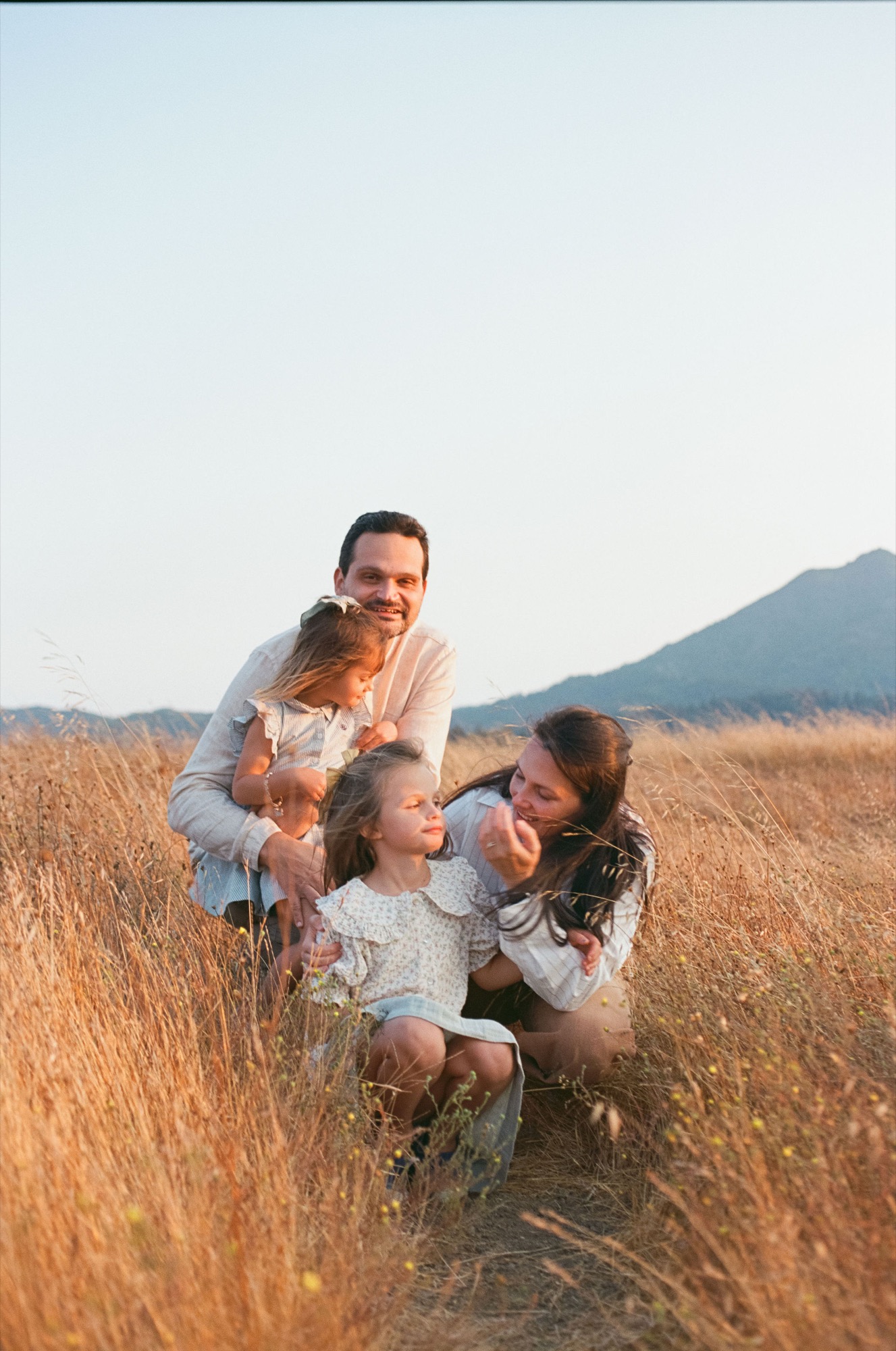 Father playfully lifts two young daughters in golden dry grass with mountains silhouetted against clear sky behind them