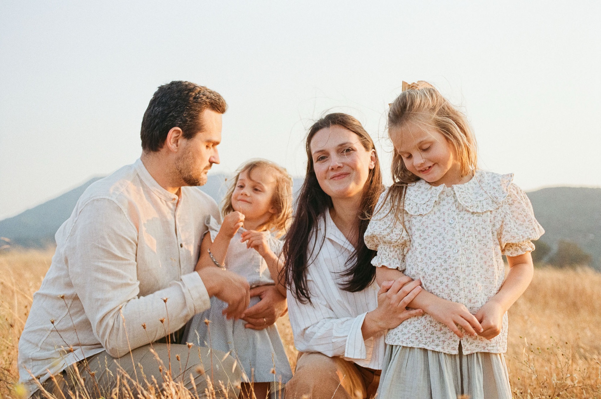 A family of four stands together in golden grassland, with parents flanking two young daughters who gaze downward with gentle smiles as afternoon light bathes the hillside