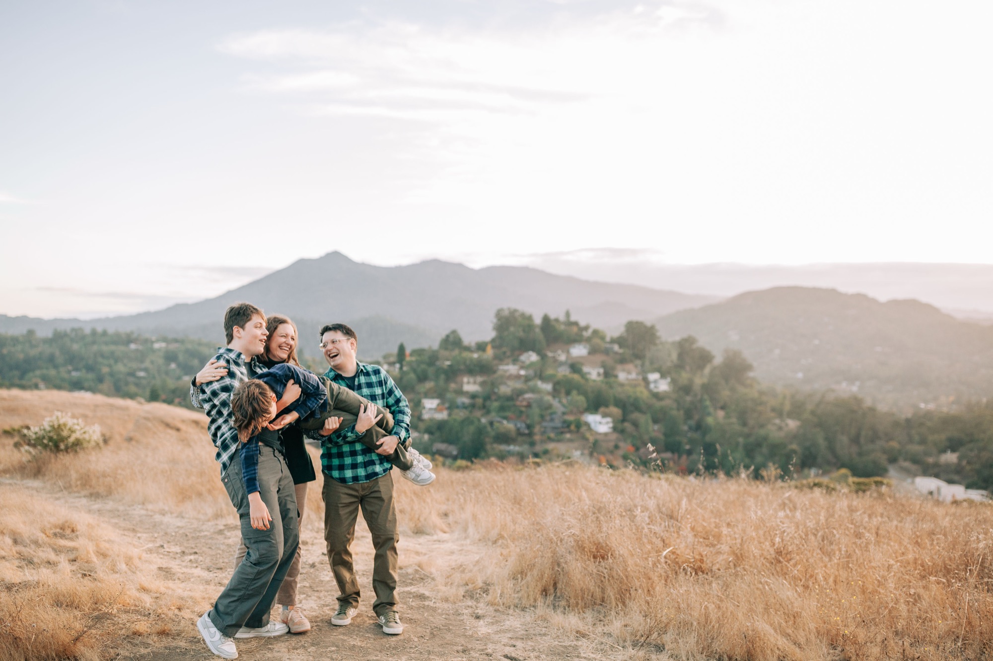 Family of four laughing as they carry their youngest sideways along a dry-grass hilltop trail, Mount Tamalpais and the San Rafael hills glowing behind them