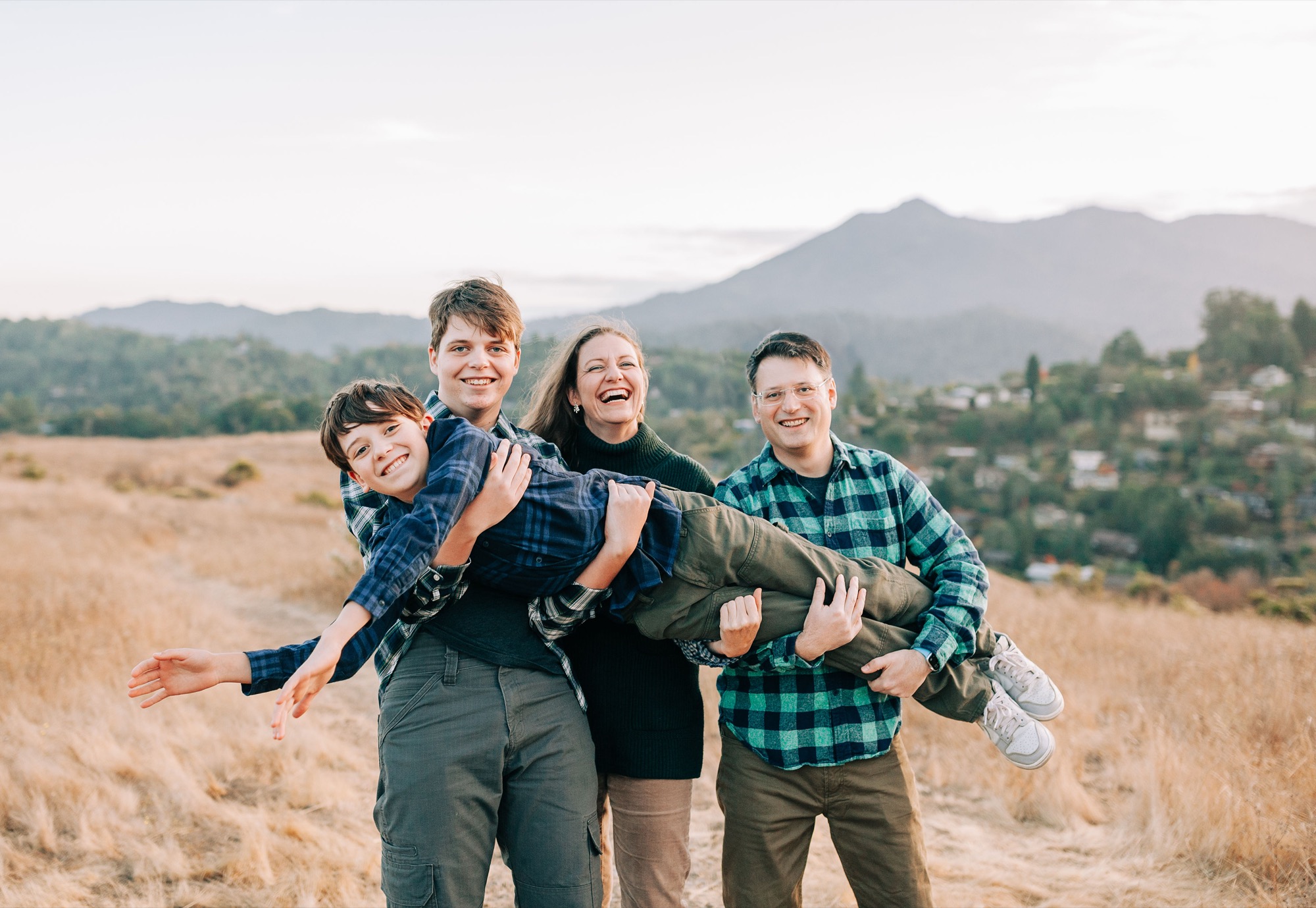 Parents and big brother hold the youngest boy flat like a plank, everyone laughing, golden grass and Mount Tamalpais in the background
