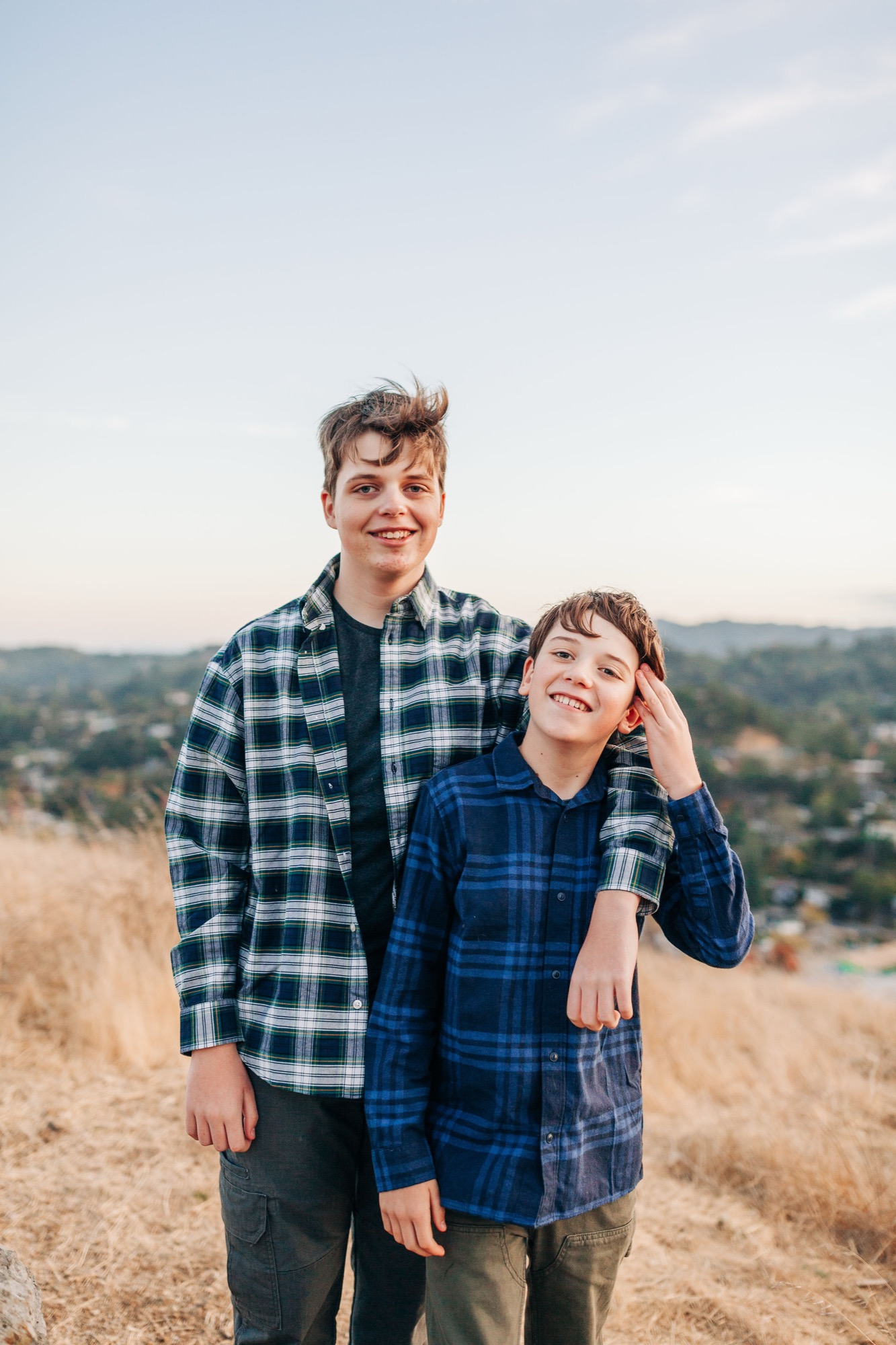 Two brothers in plaid shirts stand together on a golden hillside with the San Rafael valley soft behind them at dusk