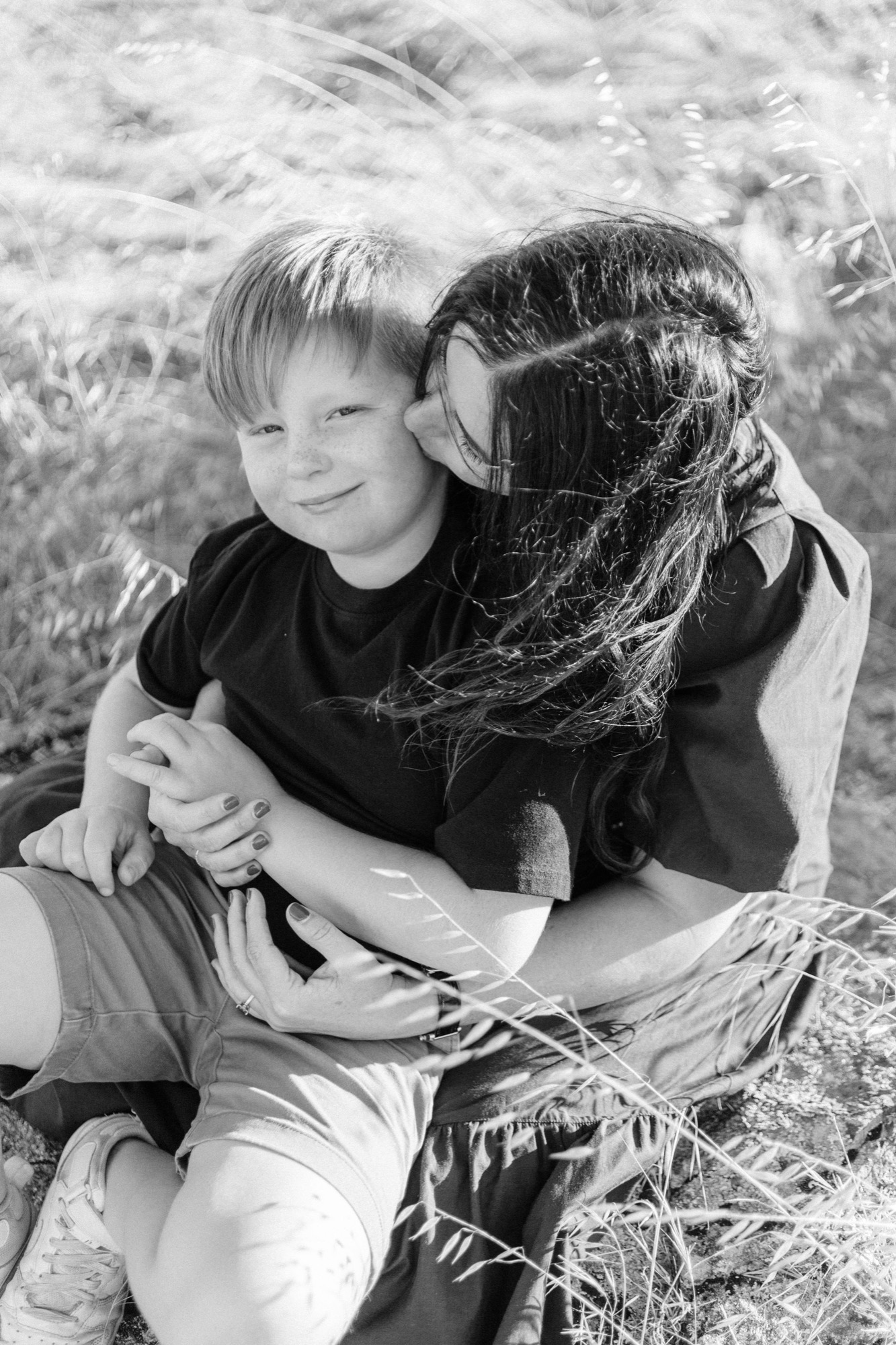 Black and white: a mom kisses her son on the cheek as they sit together in tall backlit grass