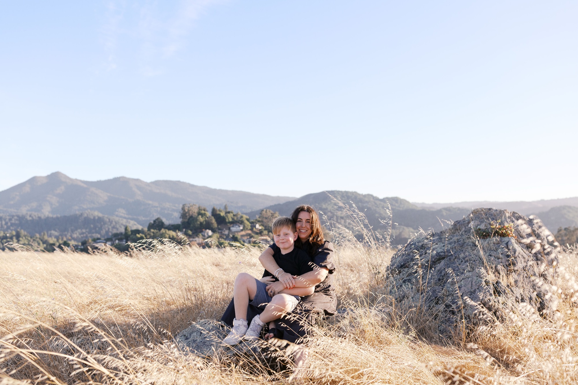 Mom and son sit on a boulder in a field of golden grass with Mount Tamalpais and the ridgeline behind them
