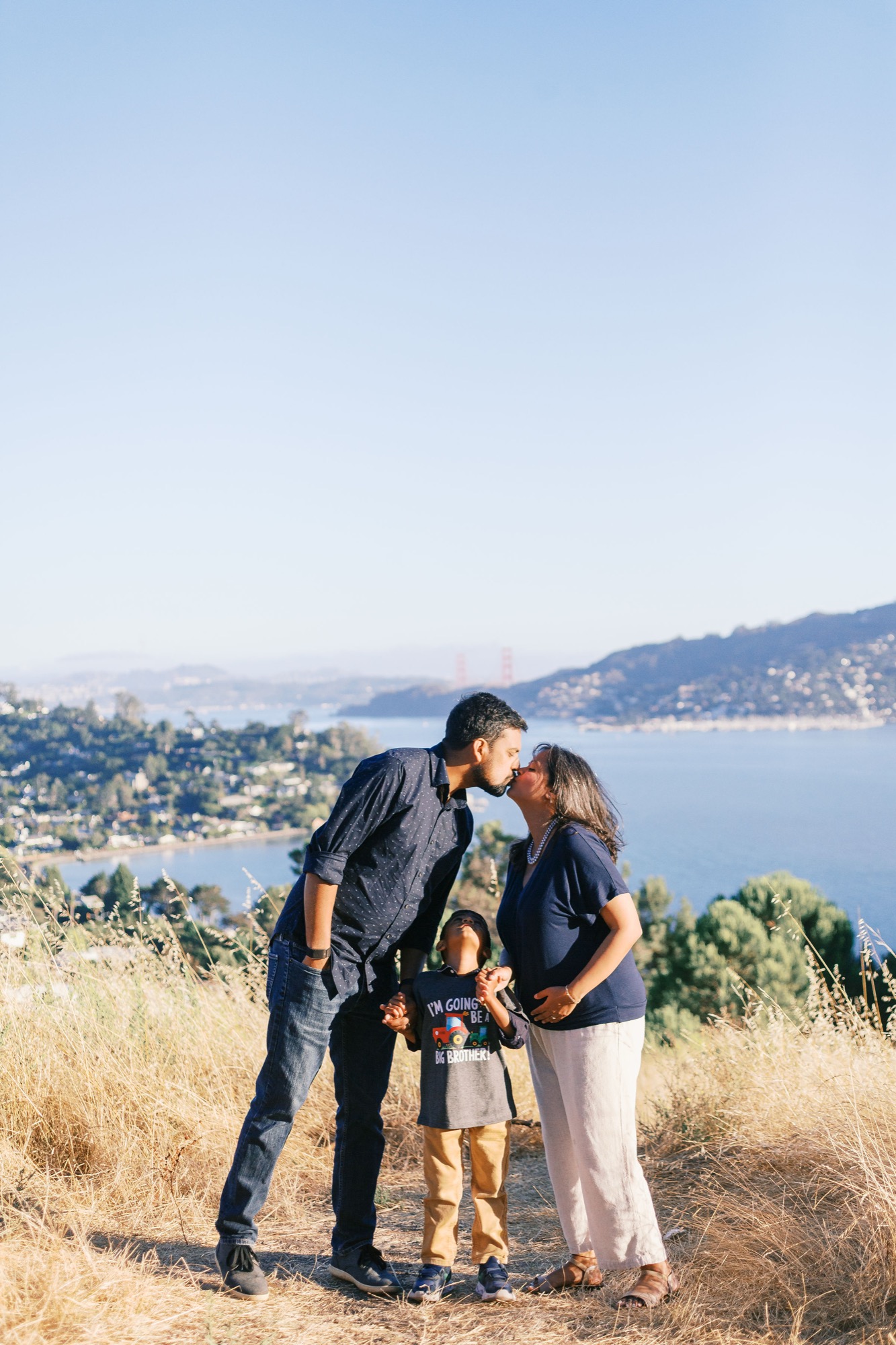 Parents kiss over their little boy's head as he looks up at them, the bay and the Golden Gate Bridge tiny in the distance