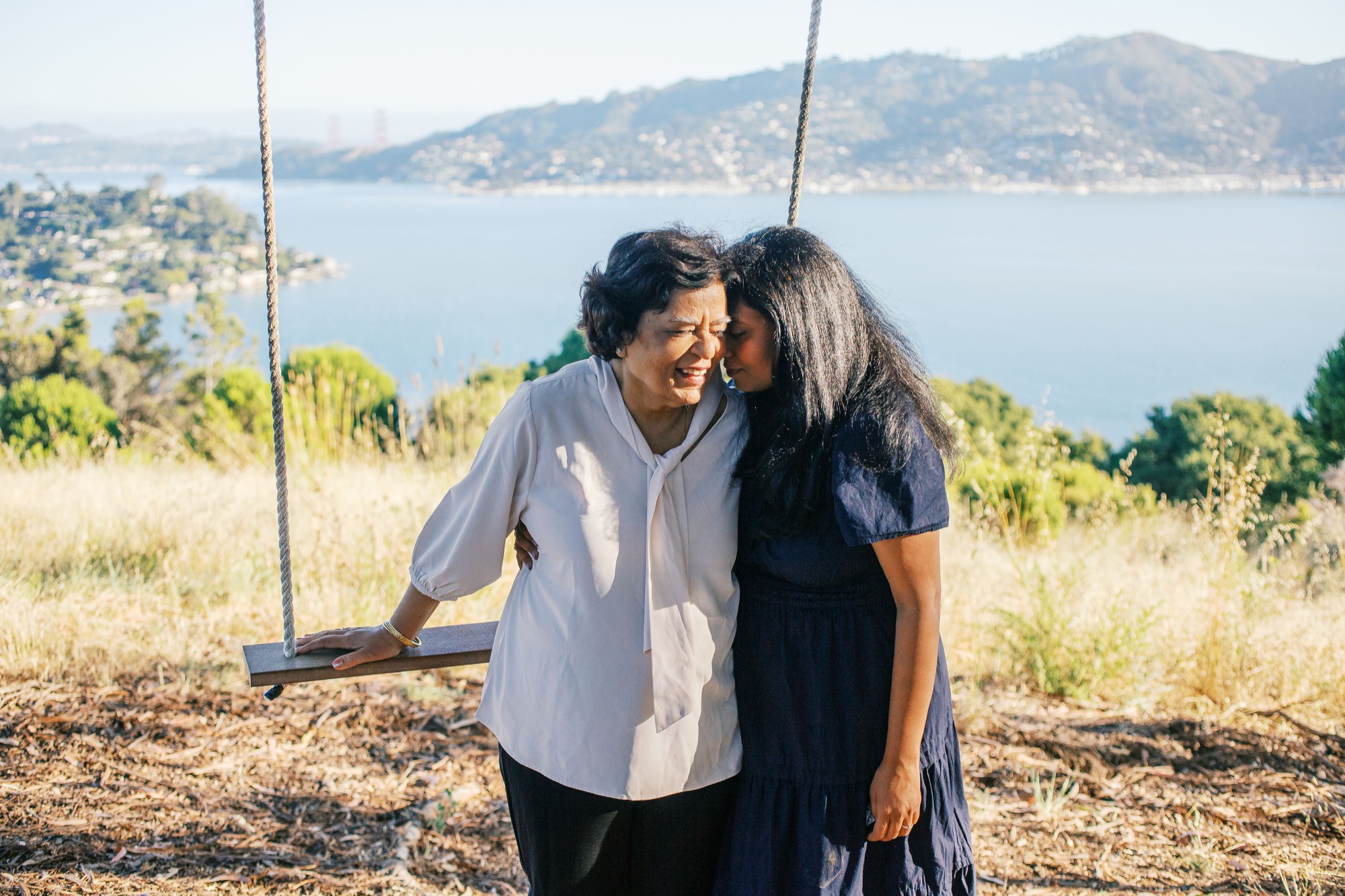 Grandmother and granddaughter forehead to forehead beside the rope swing, the bay and Tiburon hills glowing behind them