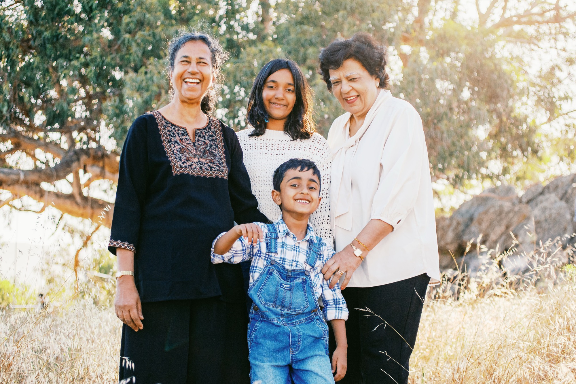Two grandmothers laugh with their grandkids in front of the eucalyptus and boulders, warm evening light behind