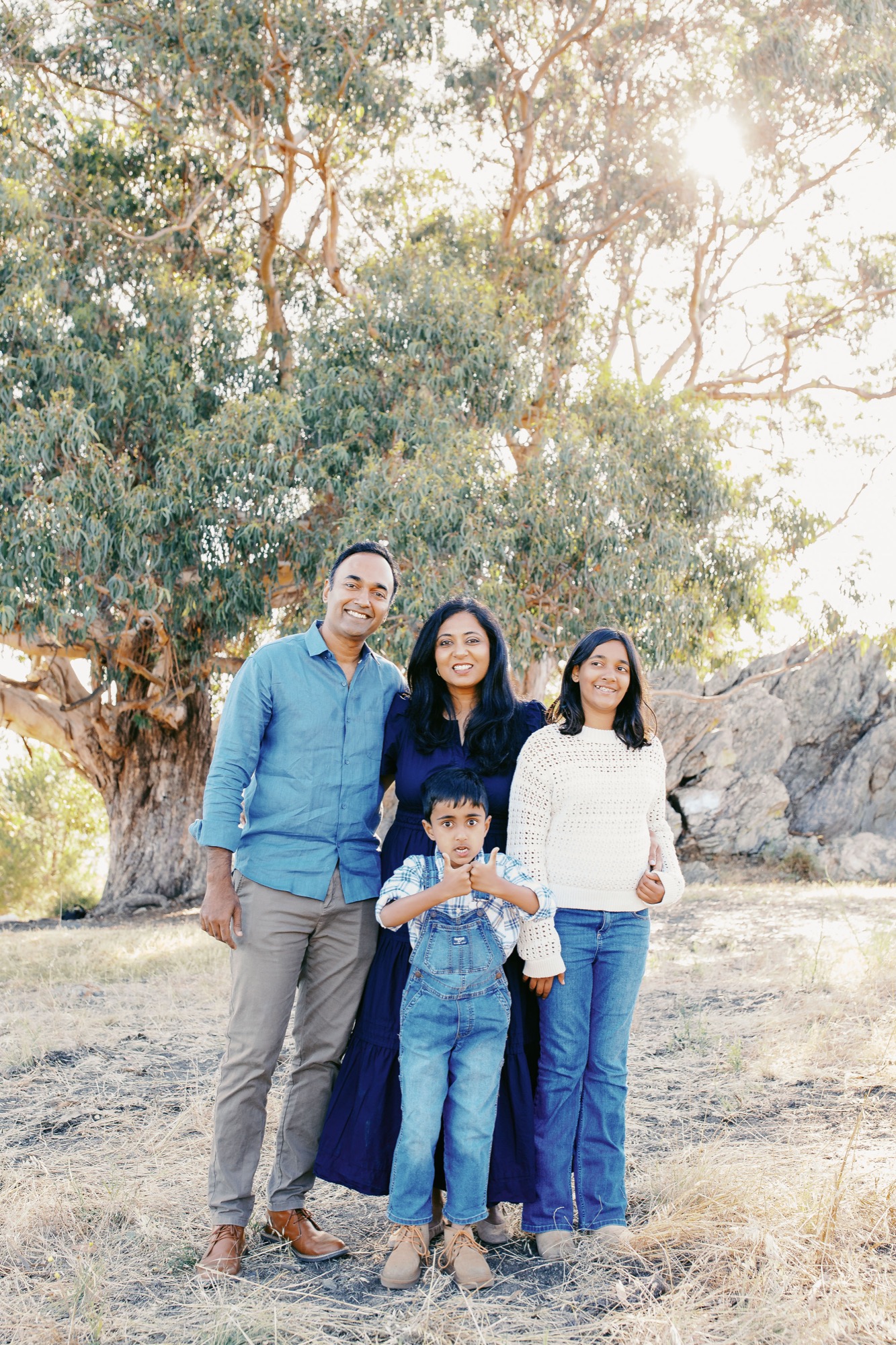 Family of four in front of the eucalyptus trunk and boulders, the little boy in overalls giving two thumbs up