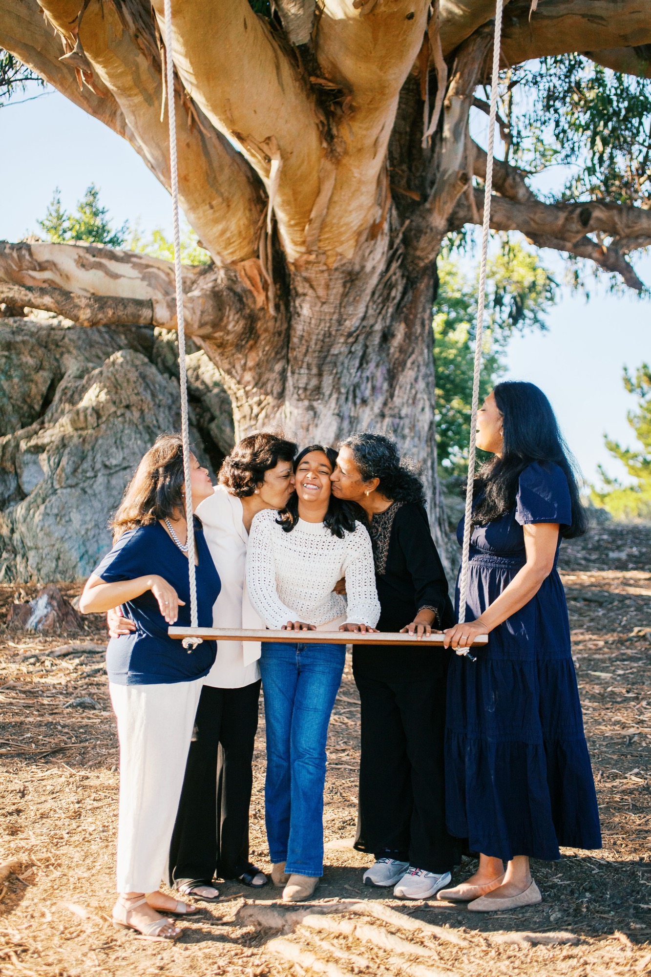 Two grandmothers and an aunt kiss a laughing girl on both cheeks at the rope swing beneath the Hippie Tree