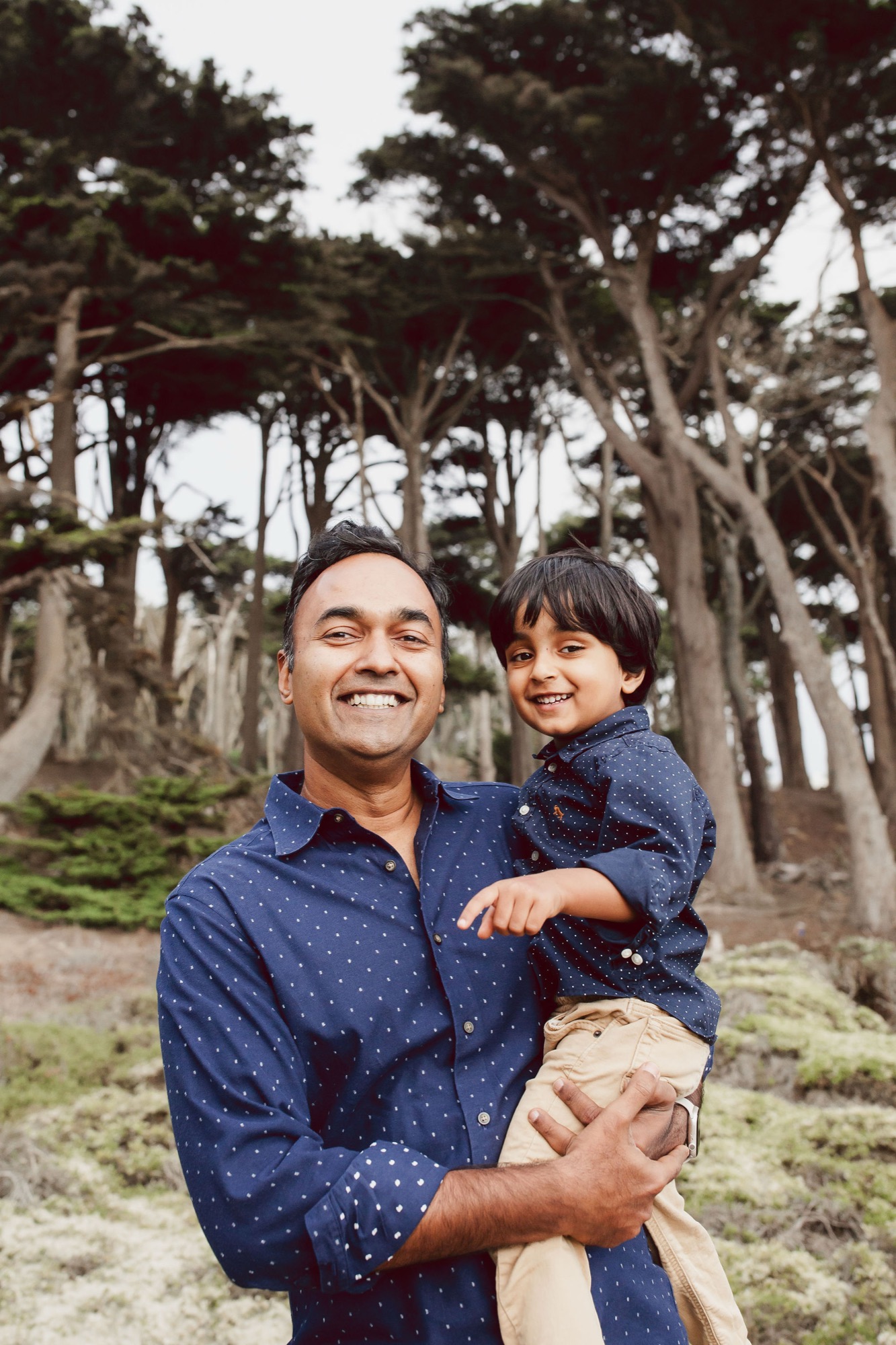 Father and young son smile broadly beneath towering Monterey cypress trees, their matching dotted shirts creating visual harmony against the natural cathedral of trunks