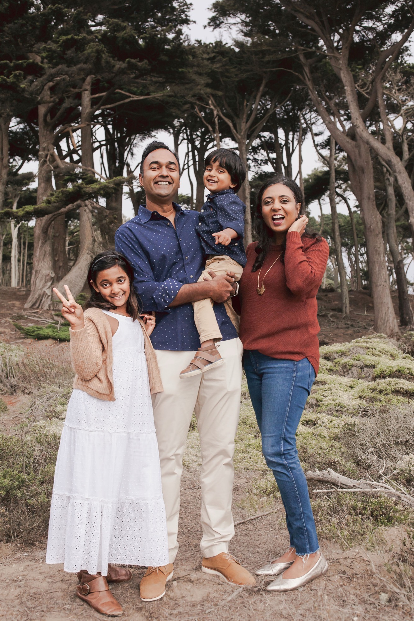 A joyful family of four stands together on a woodland path, the father holding their young son while the mother and older daughter beam at the camera beneath towering cypress trees