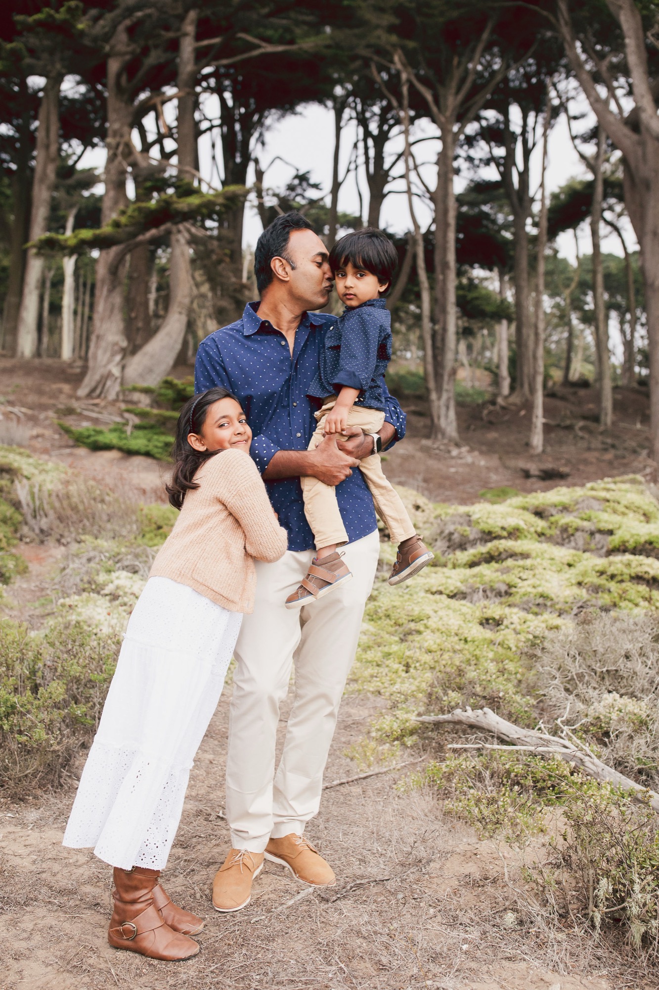 Father holds young son while daughter leans against him on a misty forest trail, all wearing coordinated blue and neutral tones