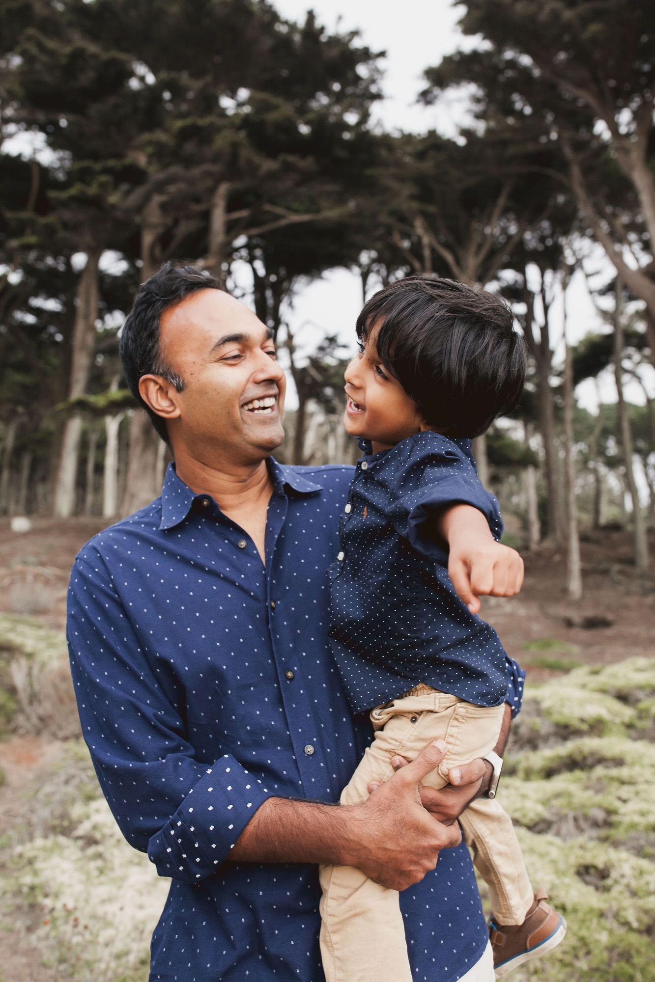 Father and young son laugh together on a tree-lined path, both wearing matching navy polka-dot shirts in soft natural light